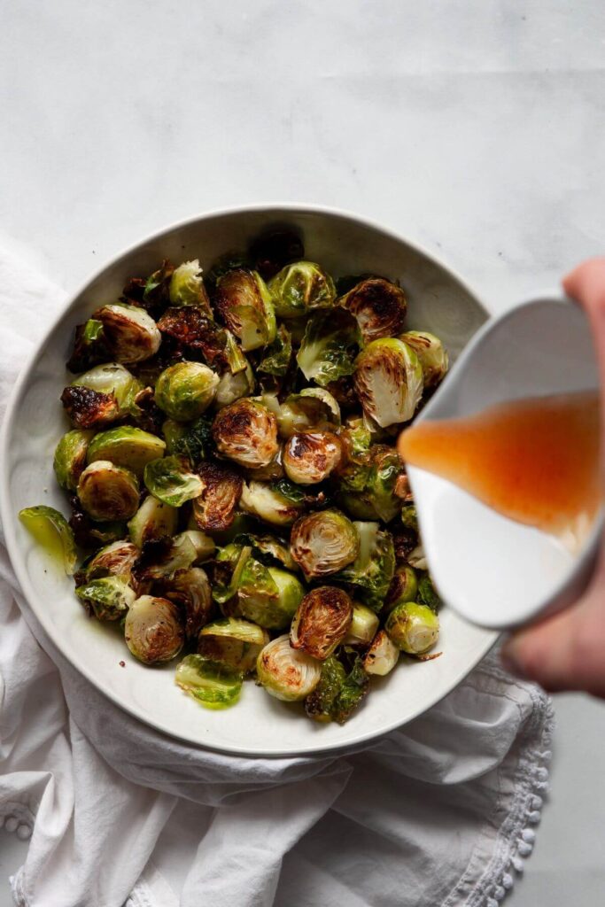pouring homemade honey sriracha sauce on top of roasted brussel sprouts in a bowl