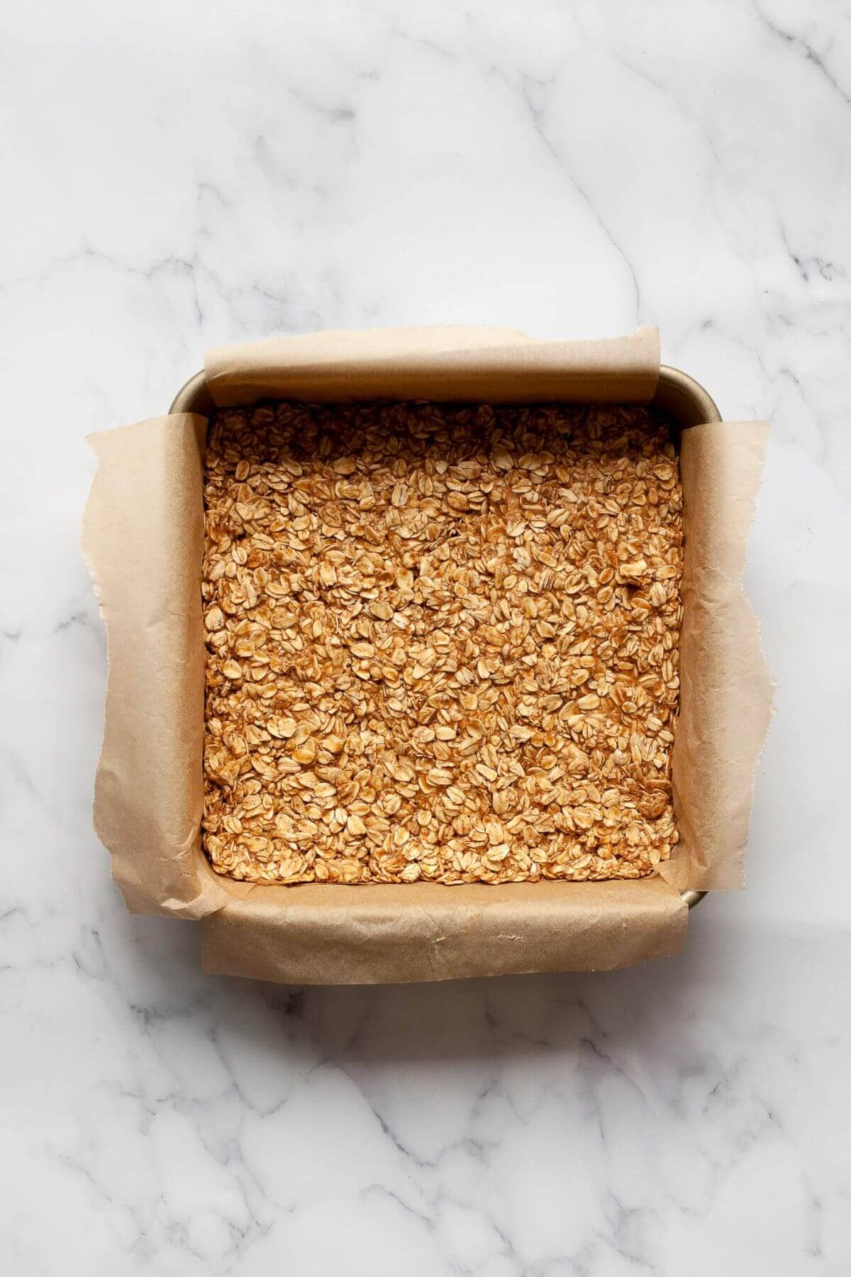 Granola base made with oats, cinnamon, vanilla, and nut butter pressed into a parchment-lined baking dish.