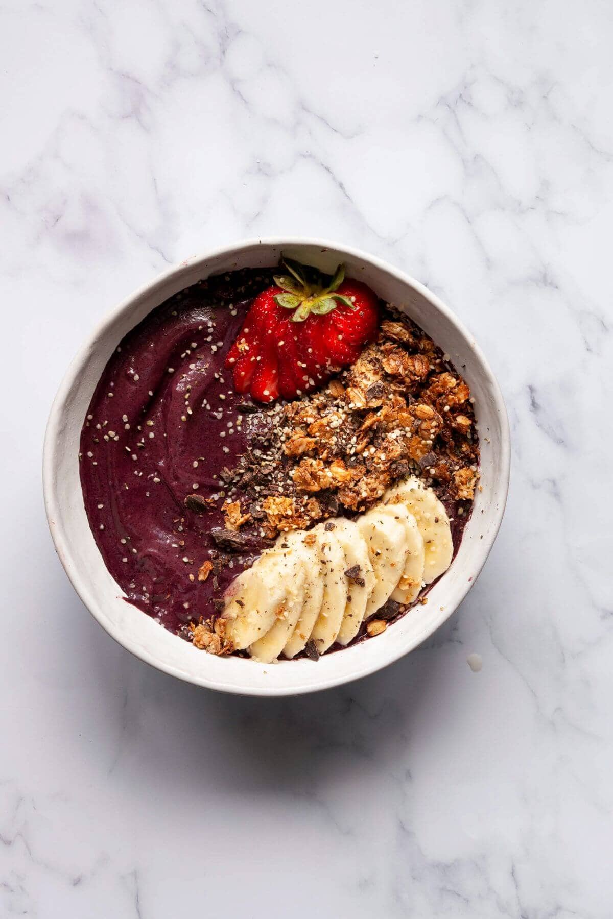 Finished high protein acai bowl in a white bowl topped with strawberries, granola, banana slices, and chia seeds.