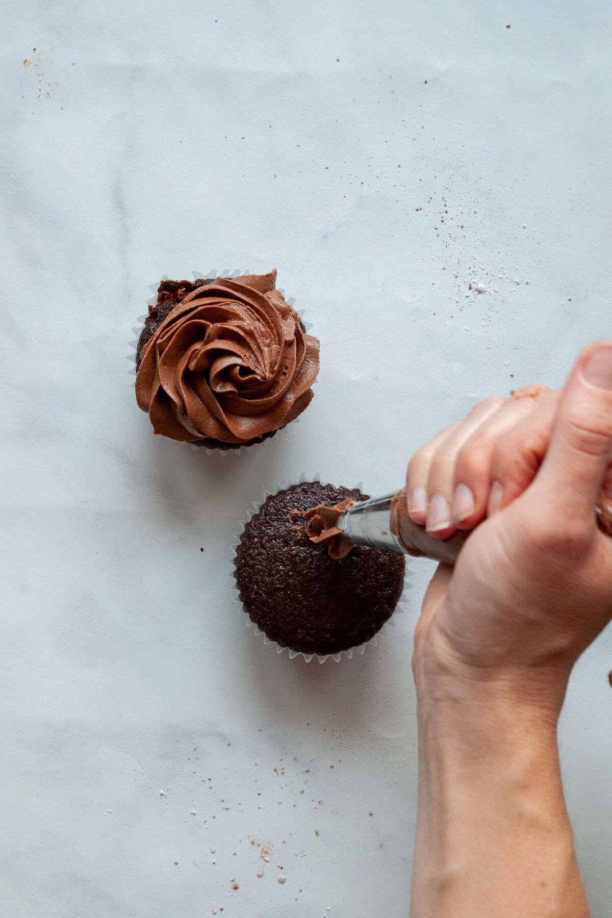 Piping dairy-free chocolate frosting onto a gluten-free chocolate cupcake by hand.