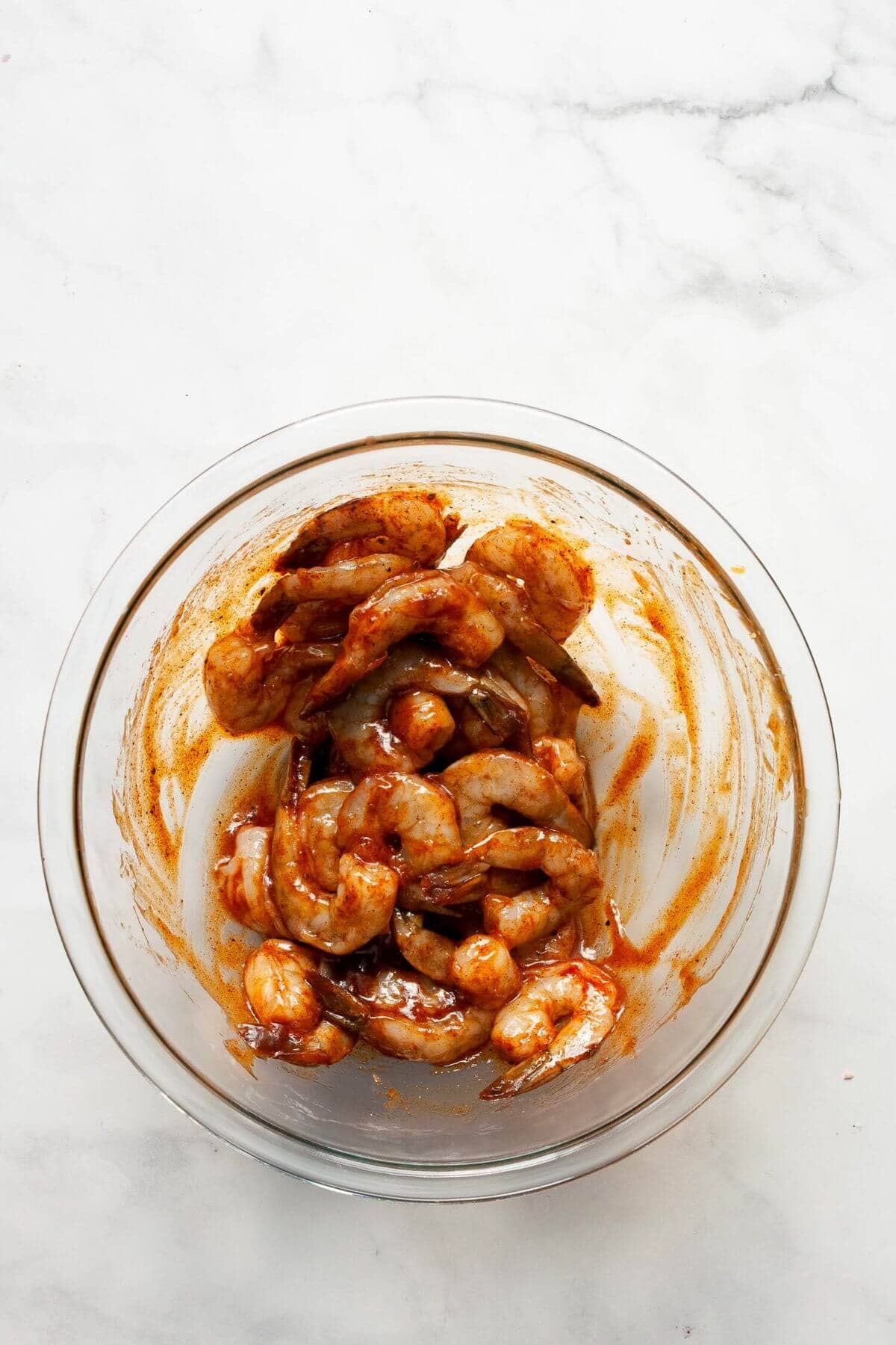 Raw shrimp coated in BBQ sauce and seasoning in a glass mixing bowl.