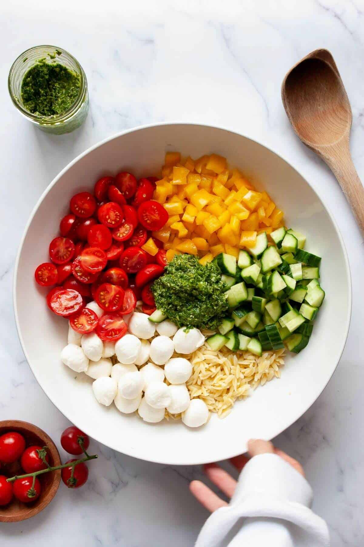 Unmixed orzo salad ingredients in a large bowl, including cooked orzo, cherry tomatoes, cucumber, bell pepper, and mozzarella balls, just before adding pesto