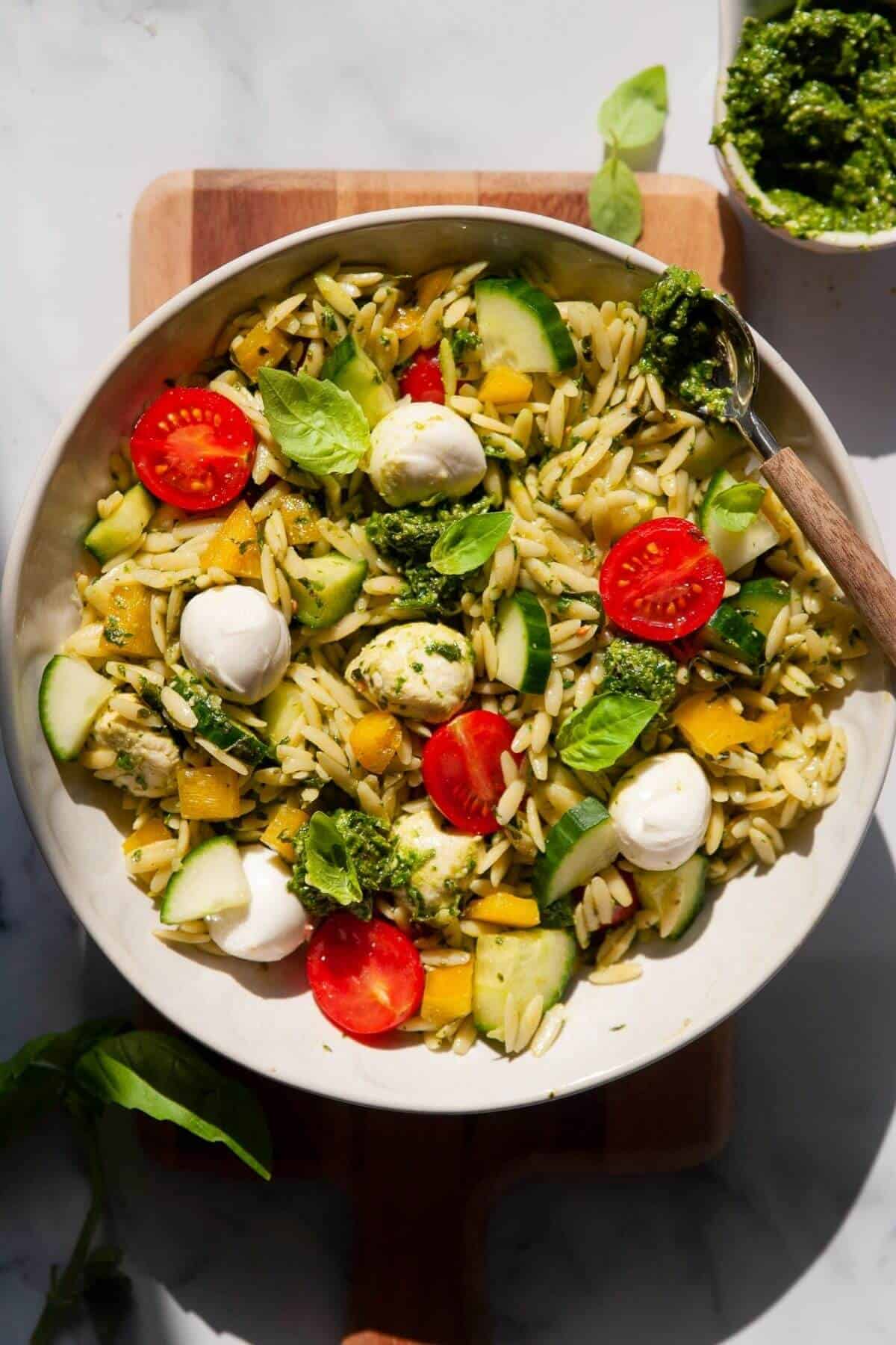 A bowl of orzo pesto salad filled with cherry tomatoes, cucumber, bell pepper, and mozzarella balls, all tossed in bright green basil pesto and garnished with fresh basil leaves