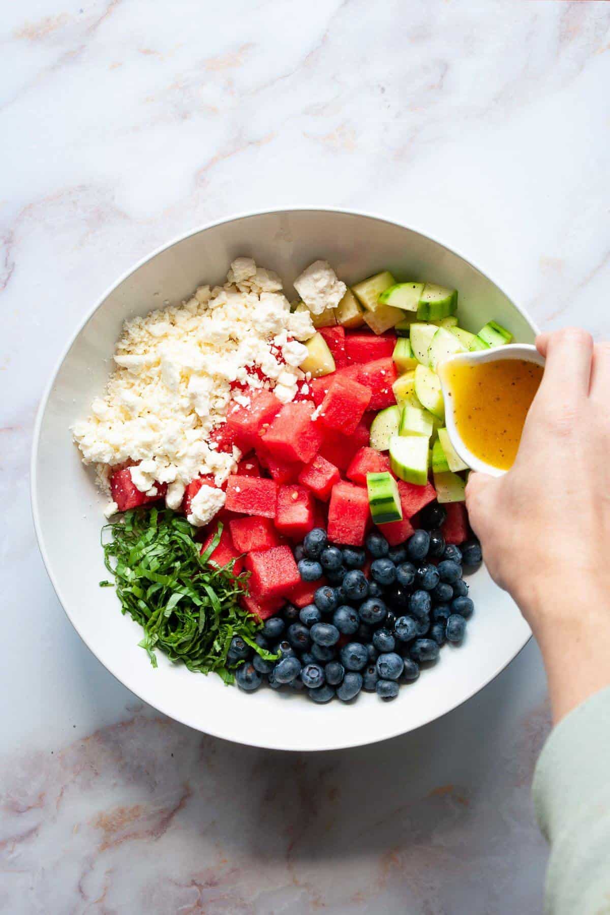 Lime dressing being poured over watermelon, cucumber, blueberries, and basil in a mixing bowl.