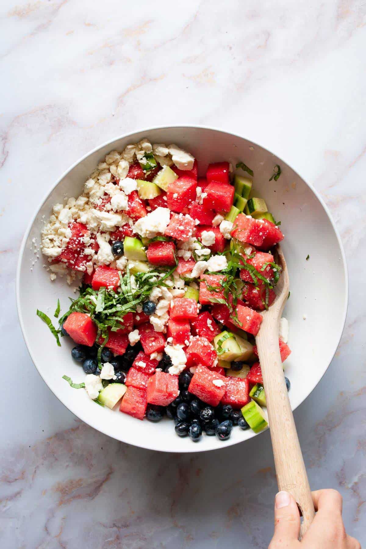 Watermelon blueberry feta salad being gently tossed together in a white bowl.