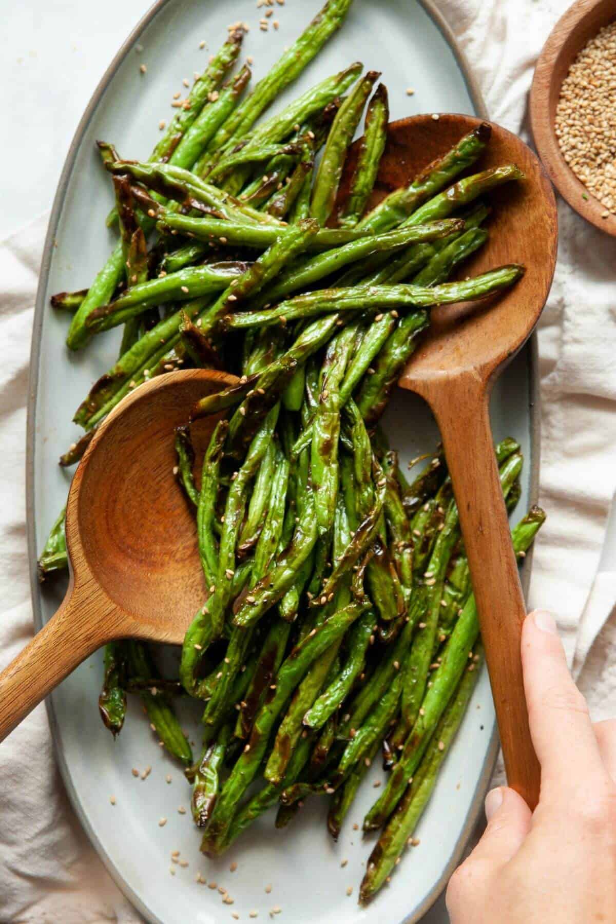 Crispy air fryer green beans topped with sesame seeds on a serving platter with two wooden spoons.