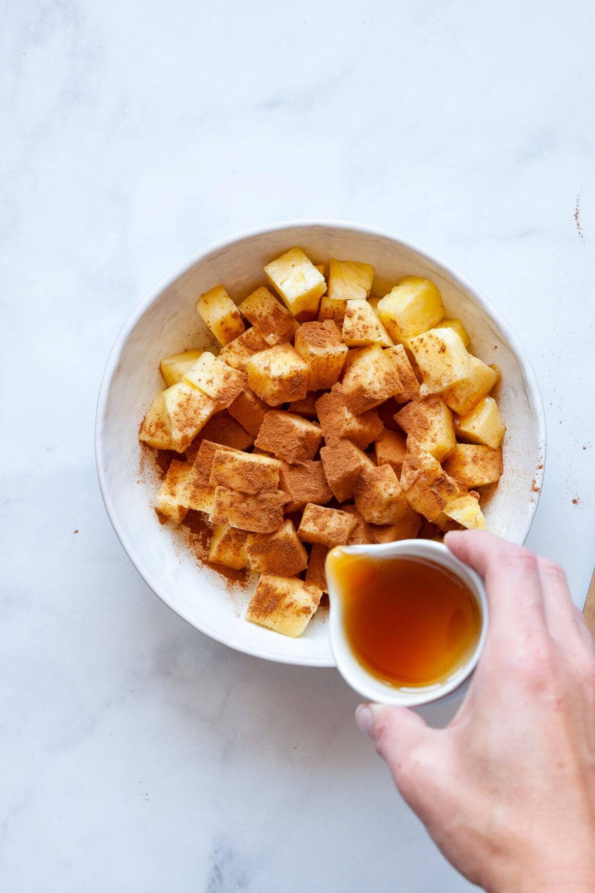 Adding cinnamon and maple syrup on top of cut up fresh pineapple in a bowl.