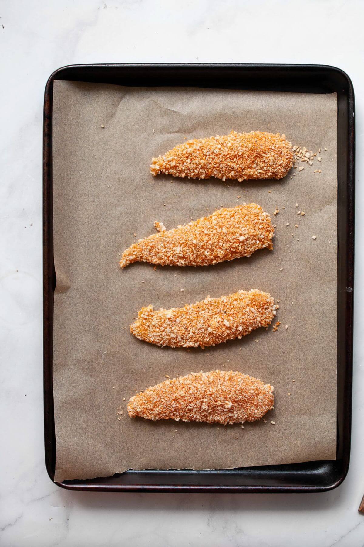 Uncooked breaded chicken tenders arranged on a parchment-lined baking sheet ready for baking.