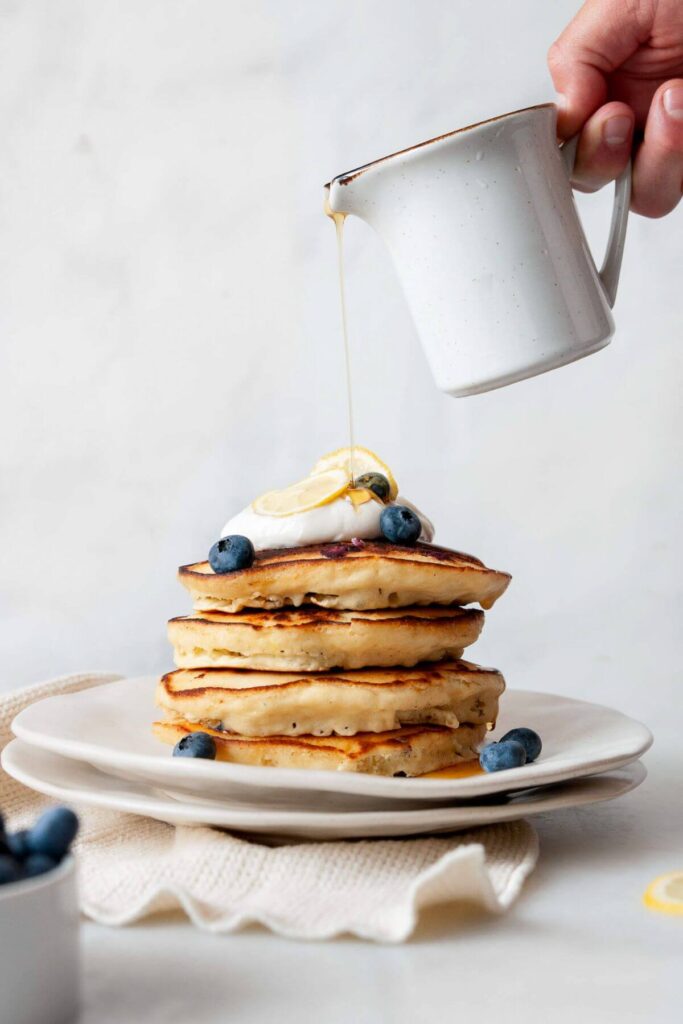 pouring maple syrup on a stack of ultra fluffy blueberry lemon pancakes