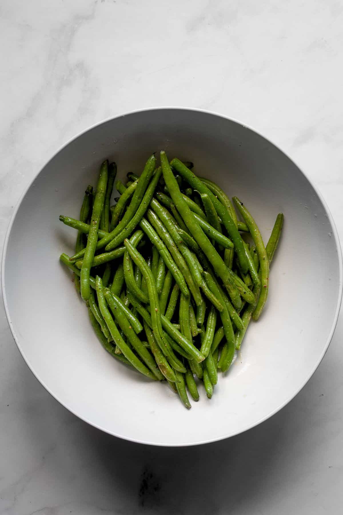 Trimmed green beans tossed with olive oil and seasonings in a white mixing bowl.
