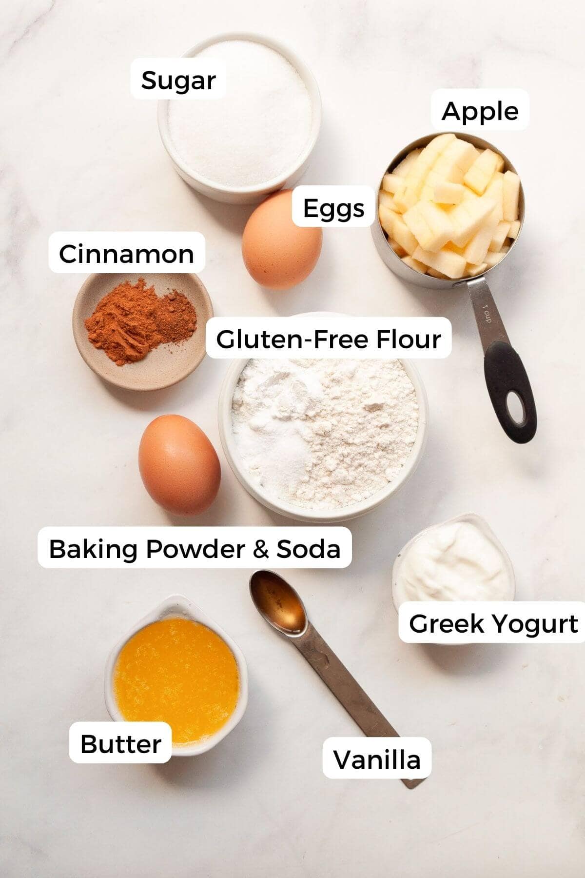 All ingredients for gluten-free apple cake in small bowls, including fresh apples, gluten-free flour blend, Greek yogurt, butter, eggs, maple syrup, cinnamon, nutmeg, and vanilla.