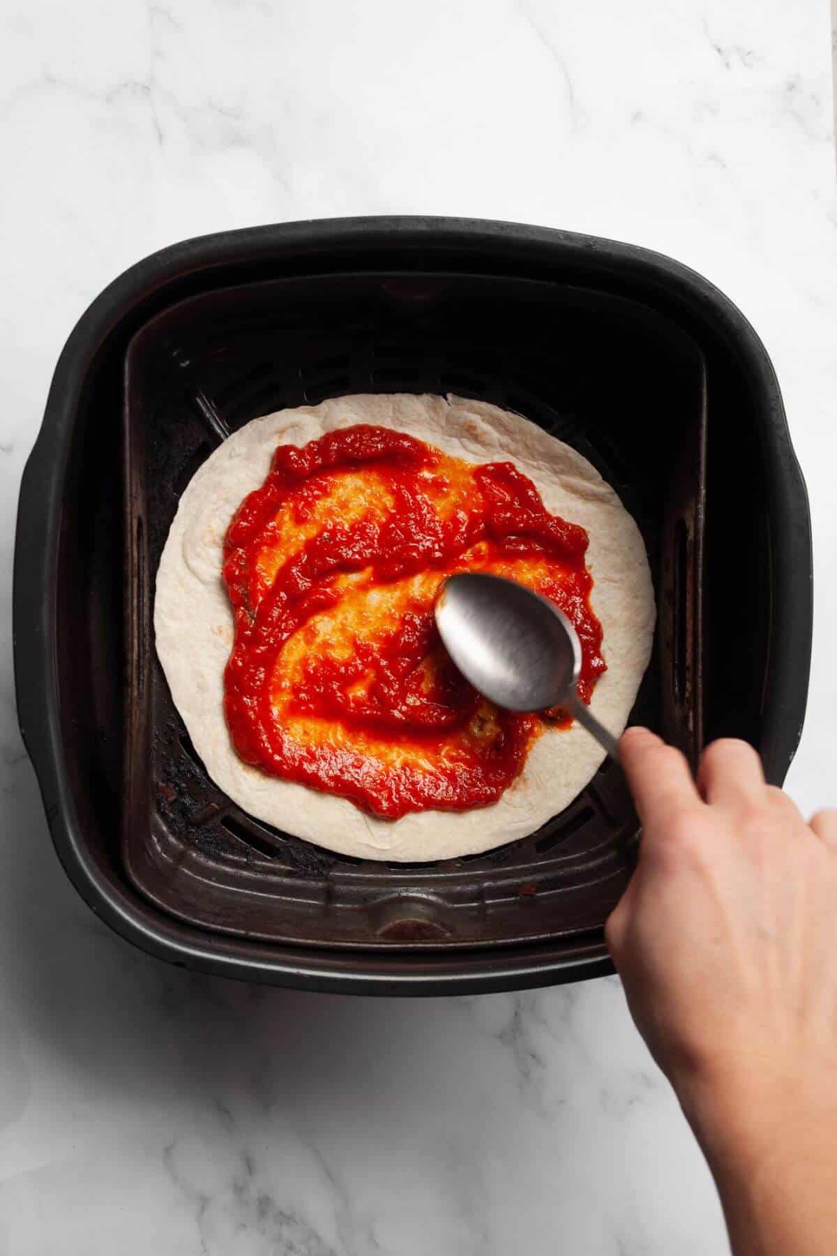Spreading pizza sauce on tortilla in air fryer basket.