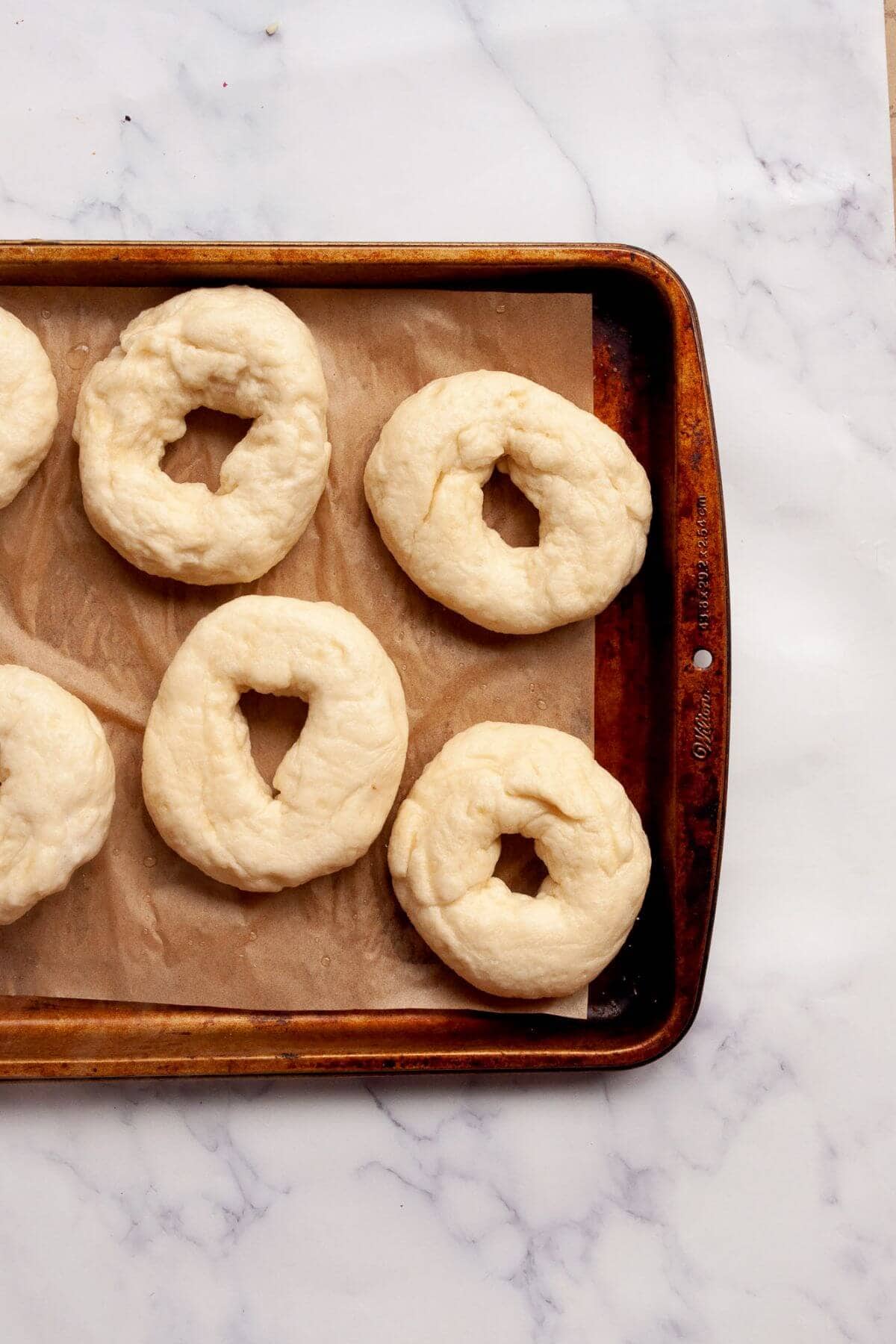 Boiled bagels with smooth, shiny surfaces on a parchment-lined baking sheet.