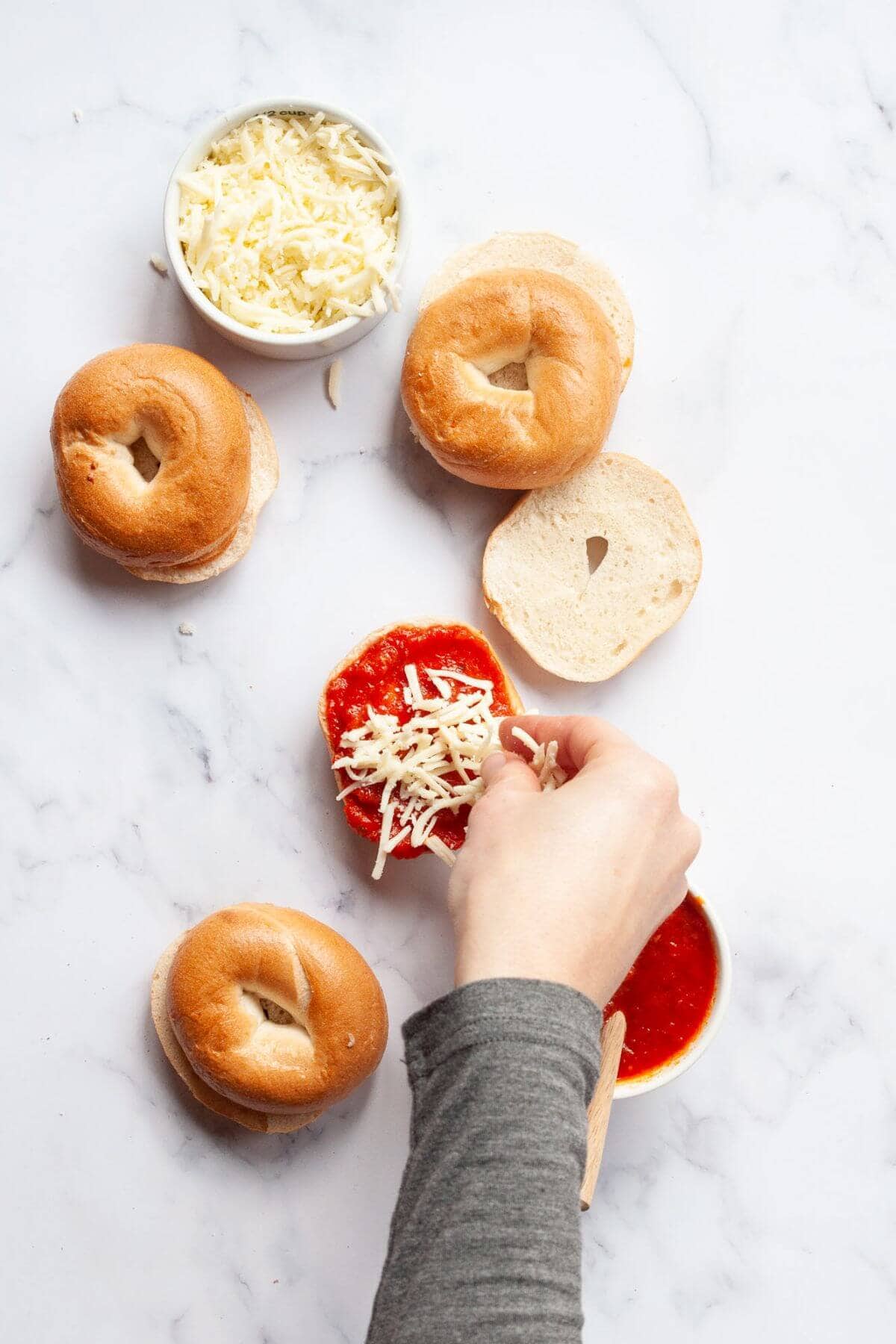 Sprinkling shredded mozzarella over pizza sauce on a mini bagel half.