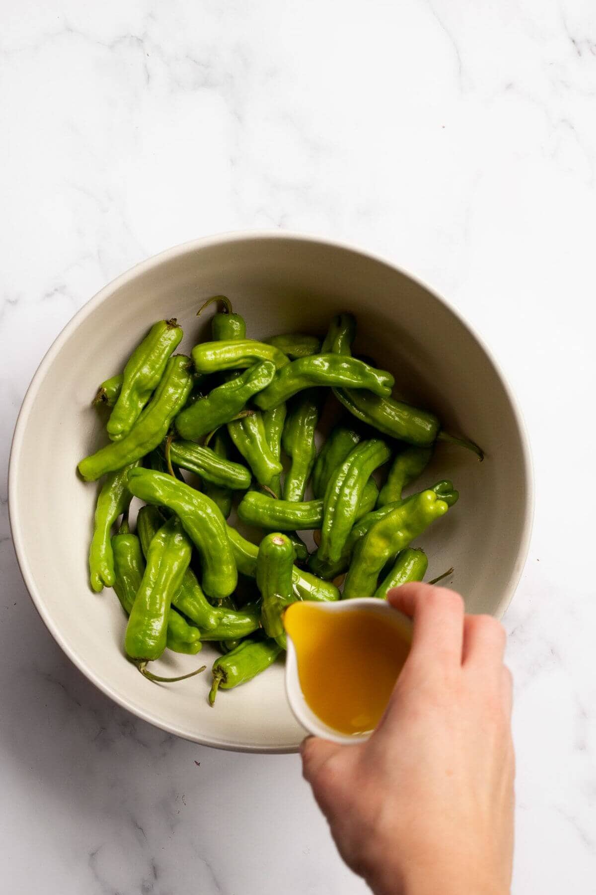 Pouring olive oil on top shishito peppers in a mixing bowl.