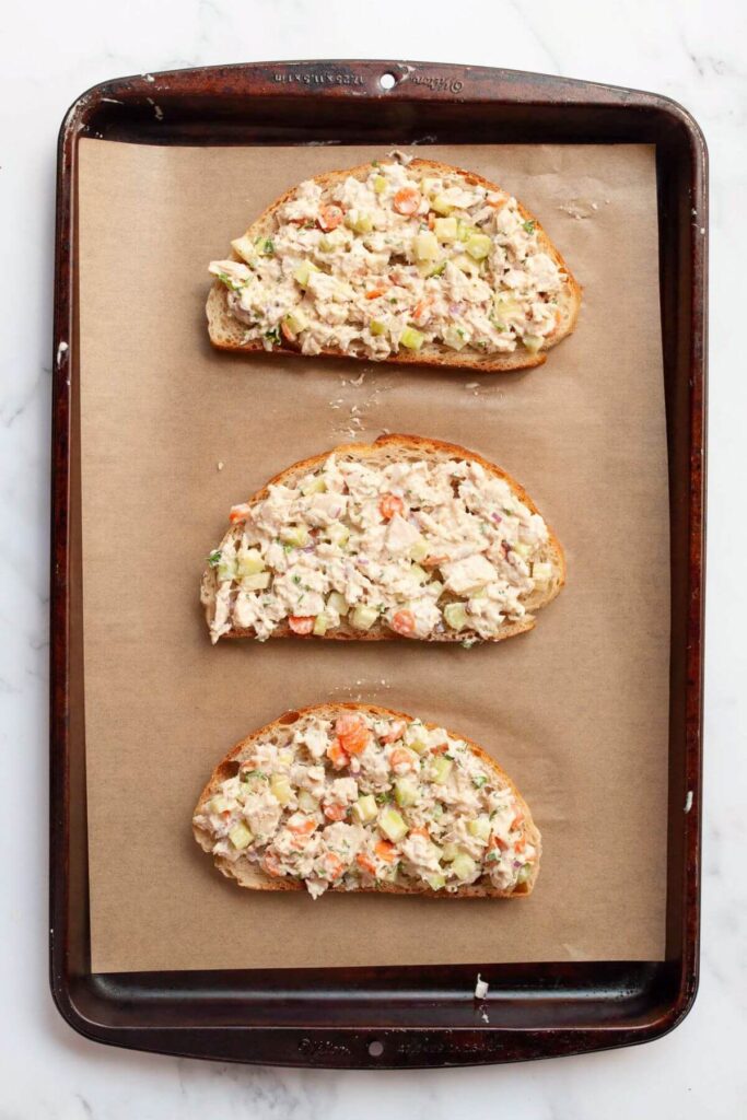 adding tuna salad on top of three slices of bread on a baking sheet