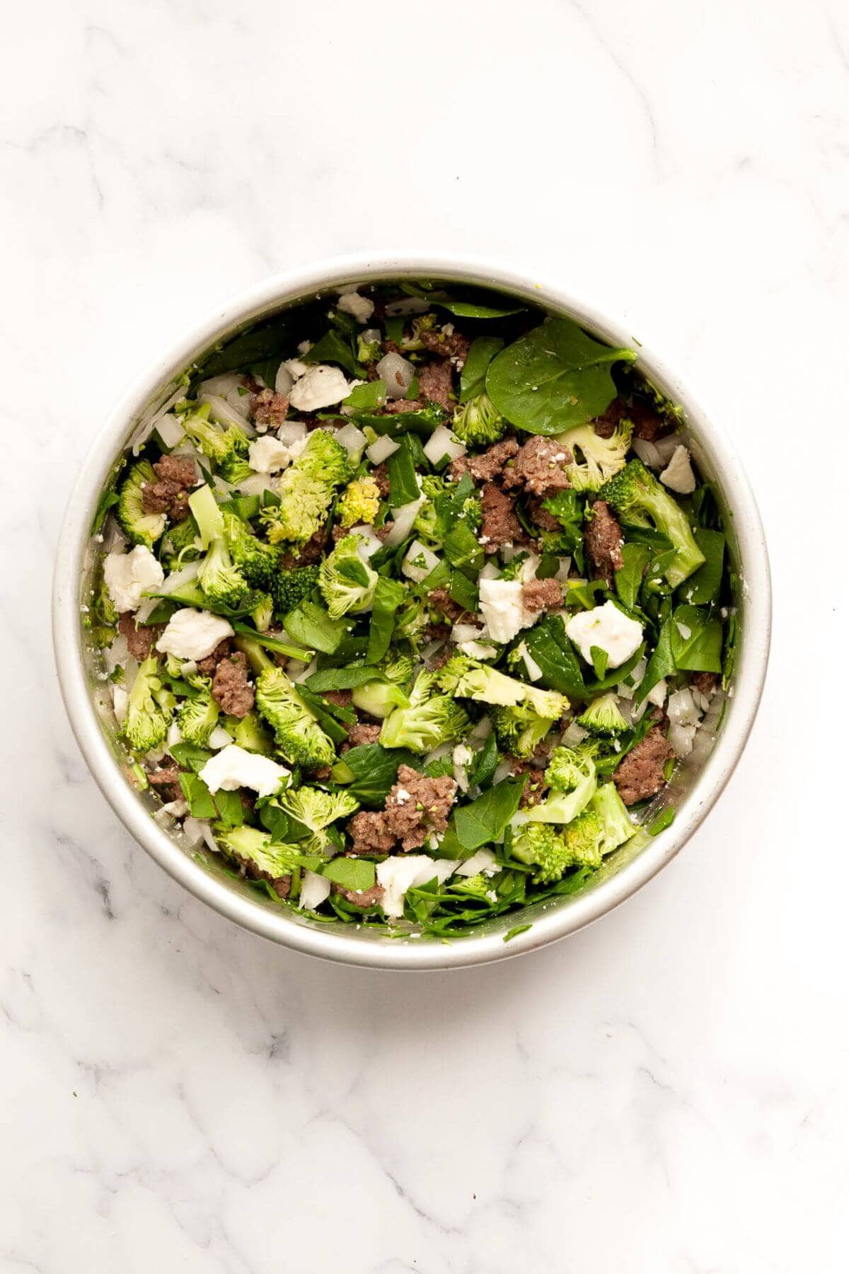 Chopped onion, broccoli, sausage, spinach, and cheese arranged in a greased air fryer pan.