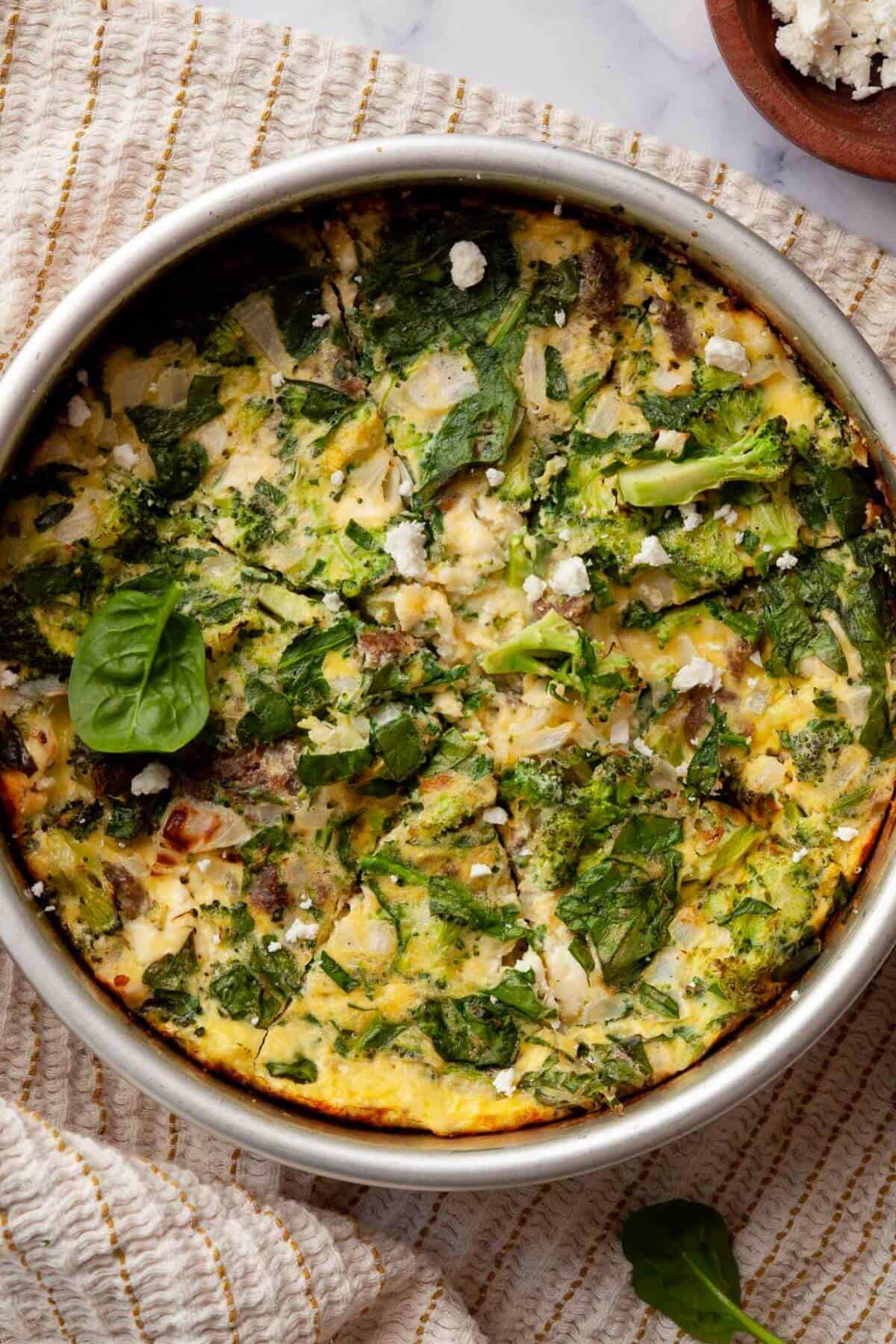 Air fryer frittata with cottage cheese in a round metal pan, topped with spinach, broccoli, and crumbled feta.