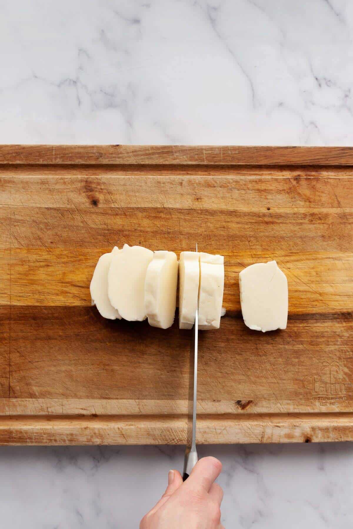 Halloumi block being sliced into half-inch thick pieces on a wooden cutting board.