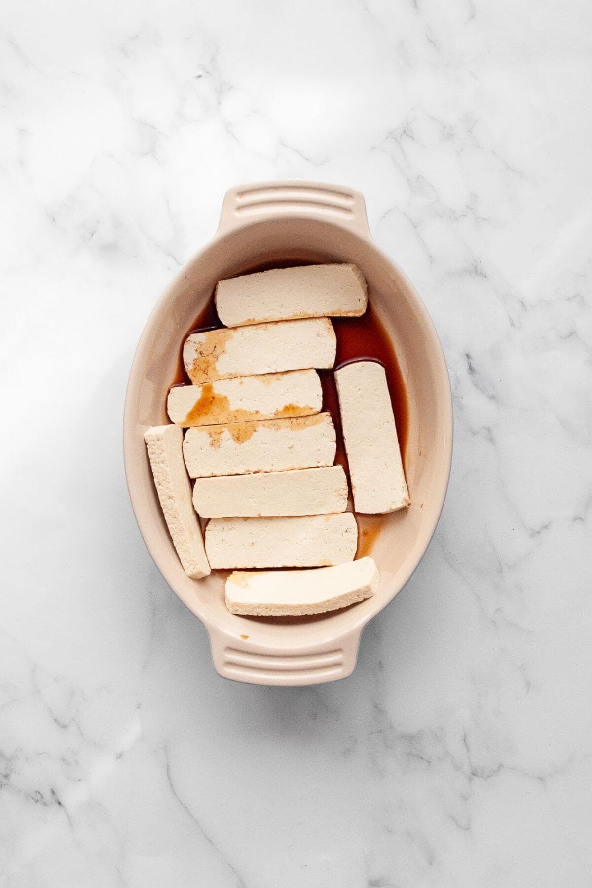 Sliced extra-firm tofu after being pressed marinating in soy sauce in a shallow dish.