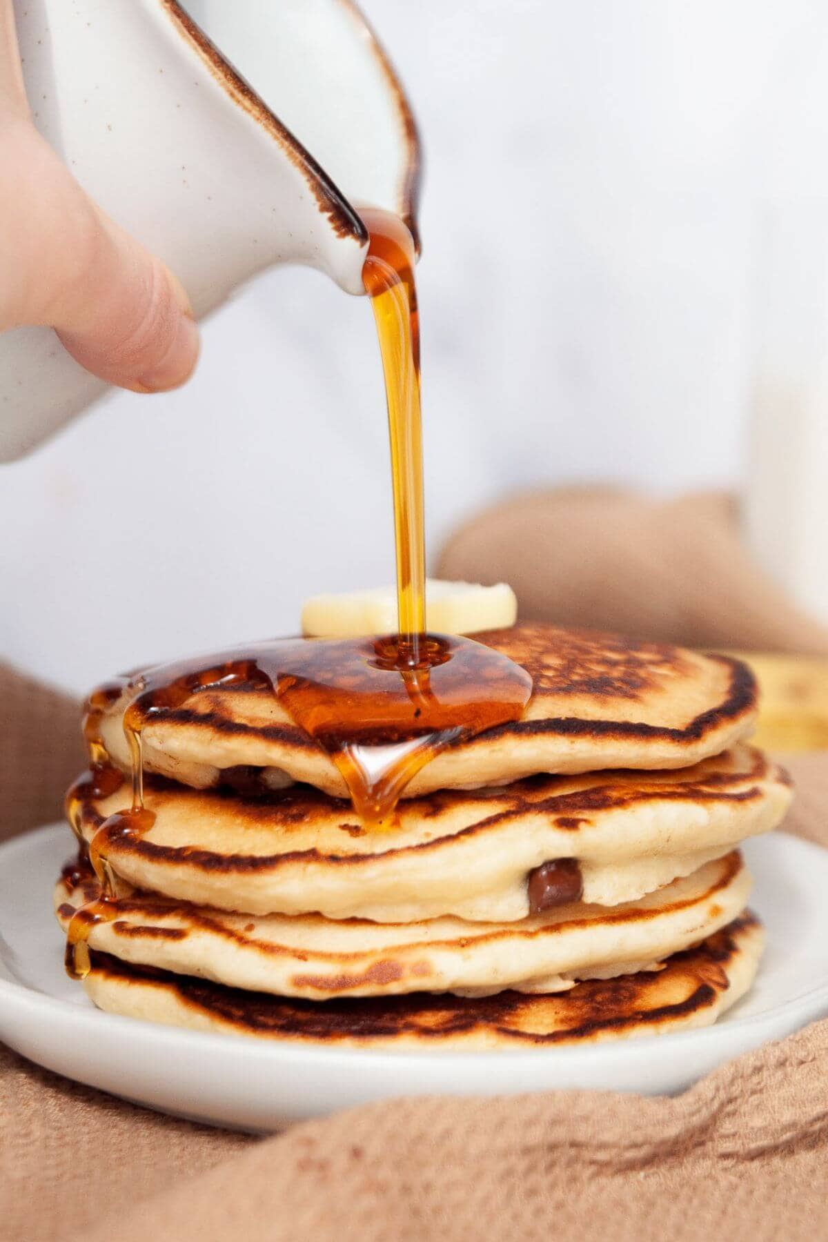 Pouring maple syrup on top of a stack of chocolate chip oat milk pancakes.