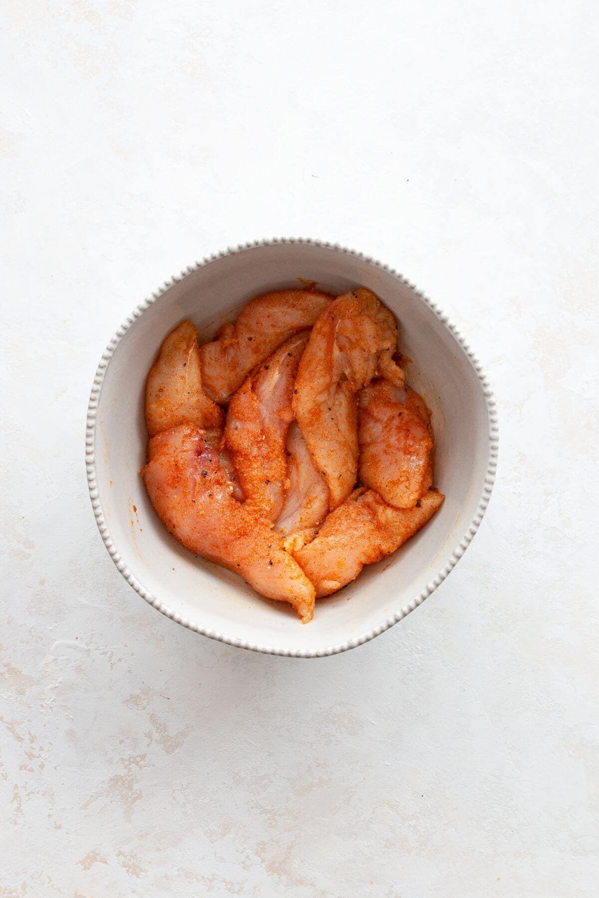 Raw chicken tenders tossed with olive oil and smoked paprika seasoning in a mixing bowl.