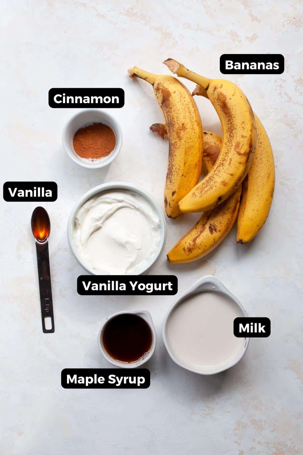 All six ingredients for banana popsicles displayed in bowls and jars, labeled as bananas, yogurt, vanilla extract, maple syrup, milk, and cinnamon.