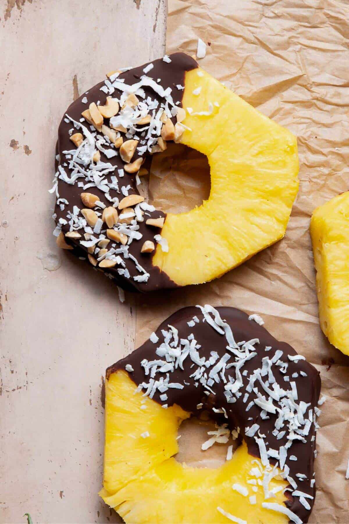 Chocolate covered pineapple rings topped with coconut and crushed nuts on brown parchment paper.