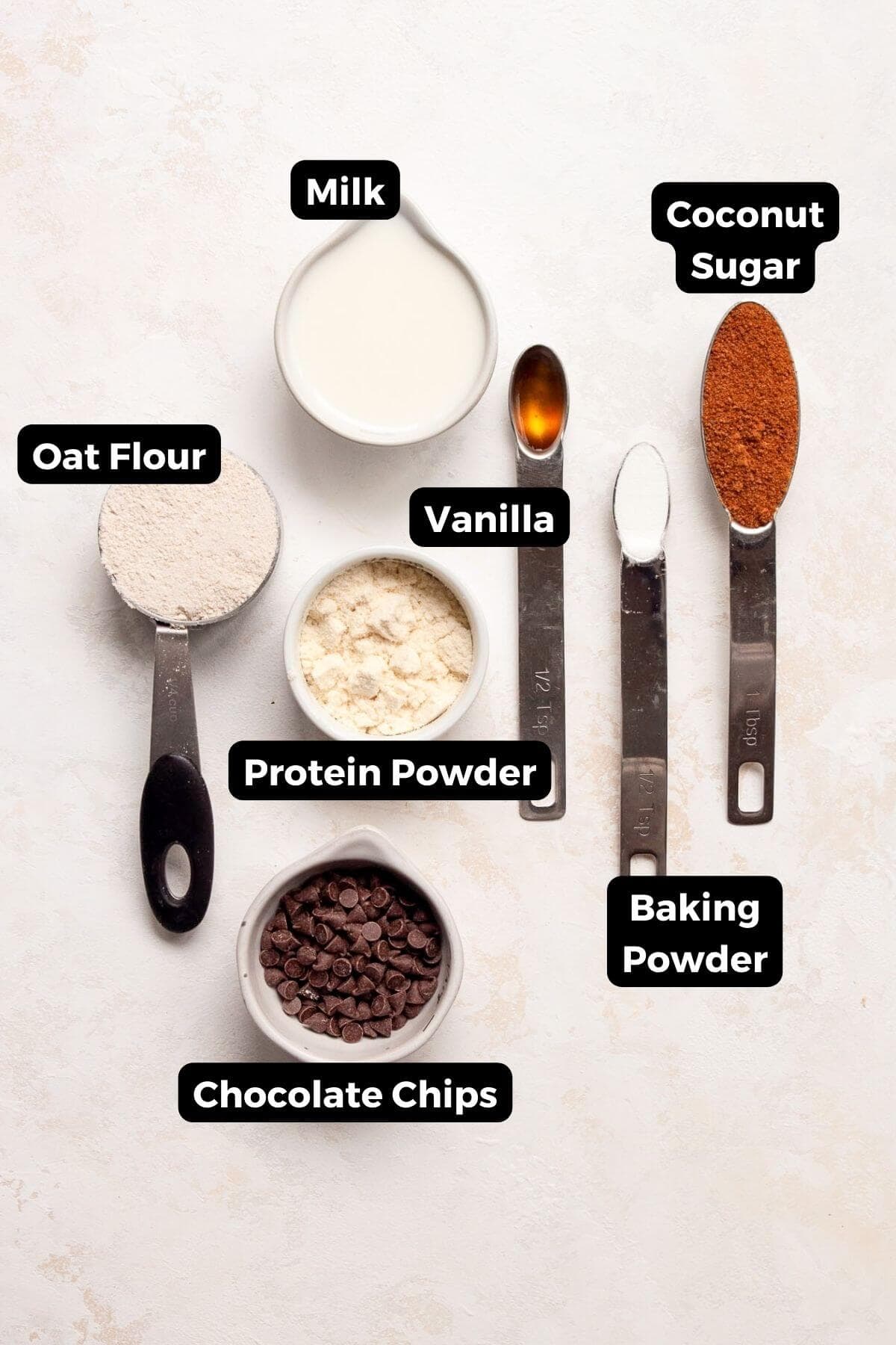 All ingredients in one minute protein mug cake recipe in small bowls including protein powder, oat flour, vanilla extract, coconut sugar, baking powder, milk, and chocolate chips.