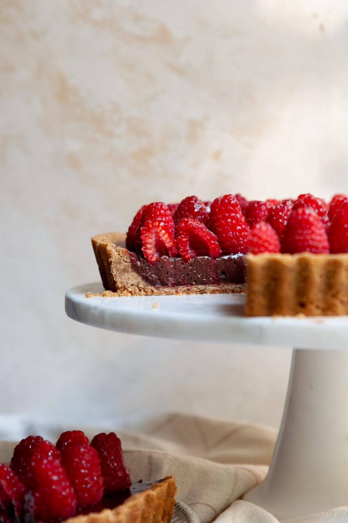 Dark chocolate raspberry tart on a white cake stand, topped with fresh raspberries and a glossy honey-jam glaze.