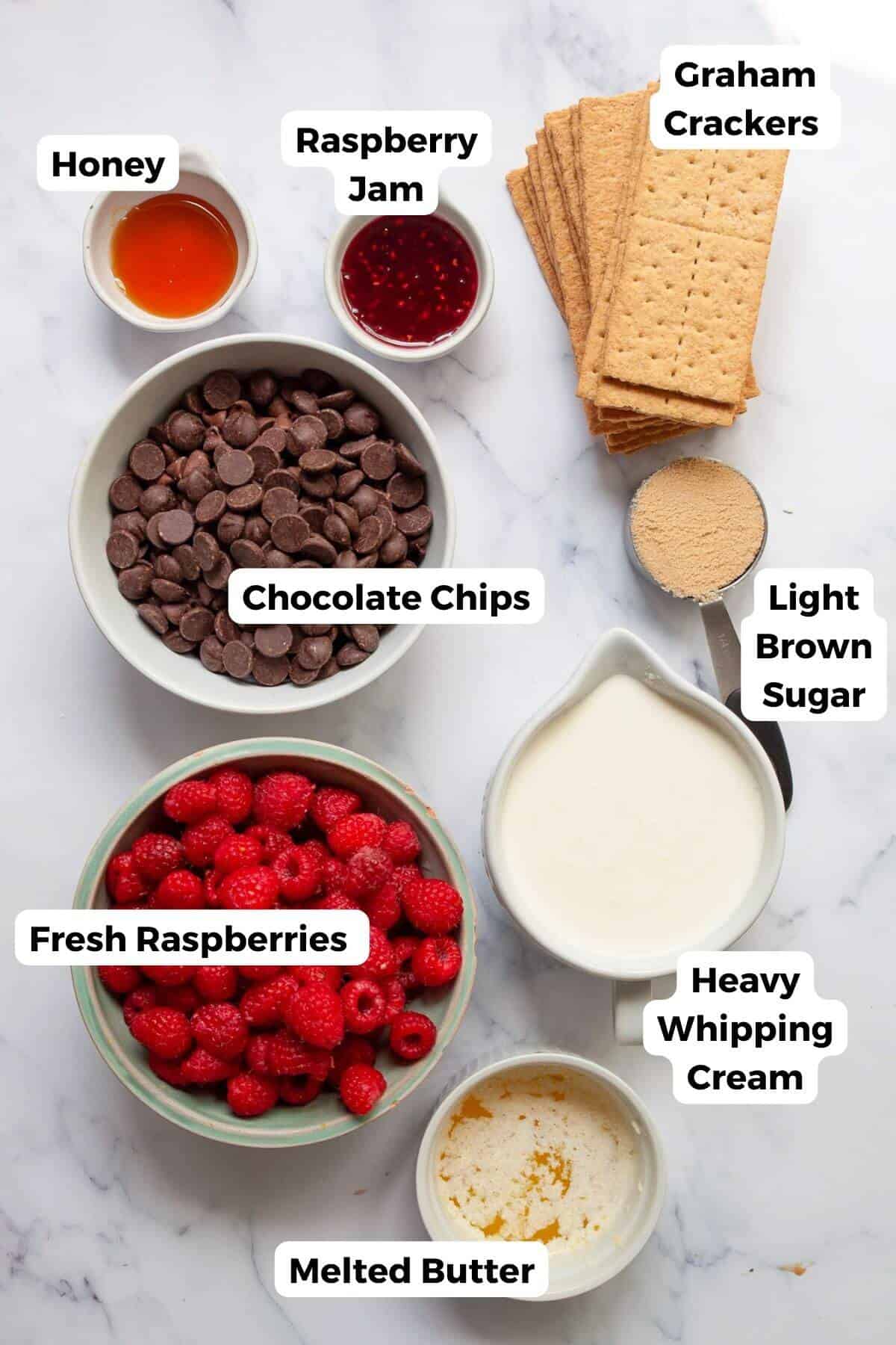 Ingredients for chocolate raspberry tart arranged on a marble surface, including graham crackers, chocolate chips, fresh raspberries, heavy cream, butter, honey, raspberry jam, and light brown sugar.