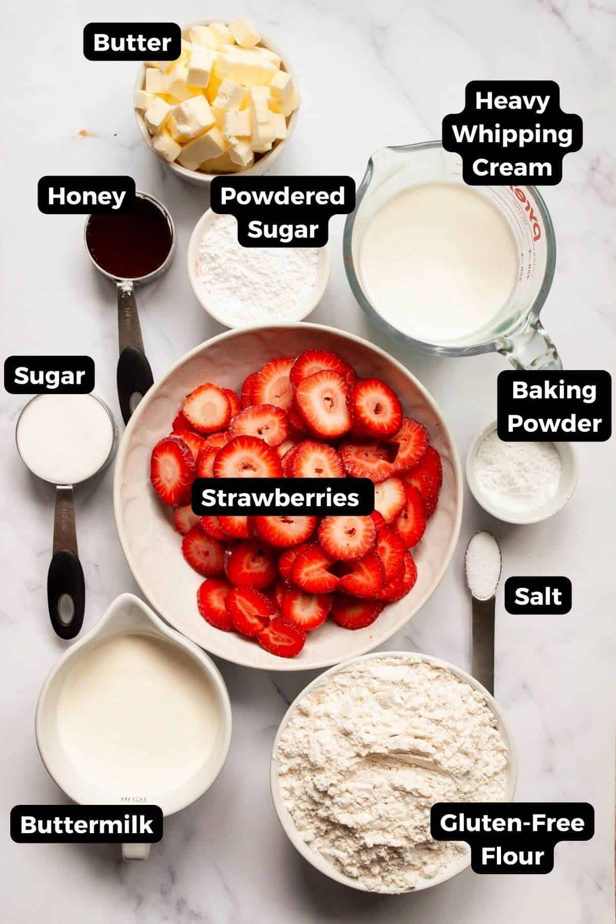 All ingredients in recipe in small bowls, including: gluten-free flour, baking powder, salt, buttermilk, fresh strawberries, honey, heavy whipping cream, and butter.