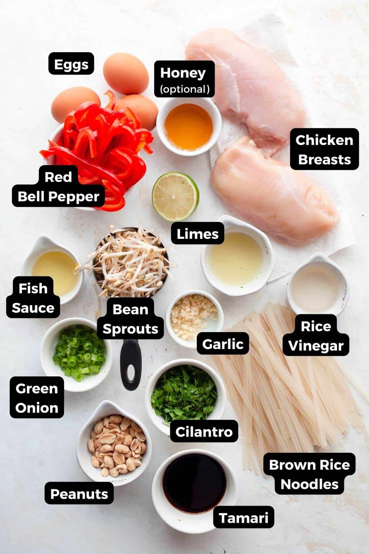 All the ingredients for healthy chicken pad Thai in small bowls with labels, including rice noodles, sliced chicken breast, eggs, carrots, bell pepper, scallions, garlic, tamari, fish sauce, lime, cilantro, and peanuts.