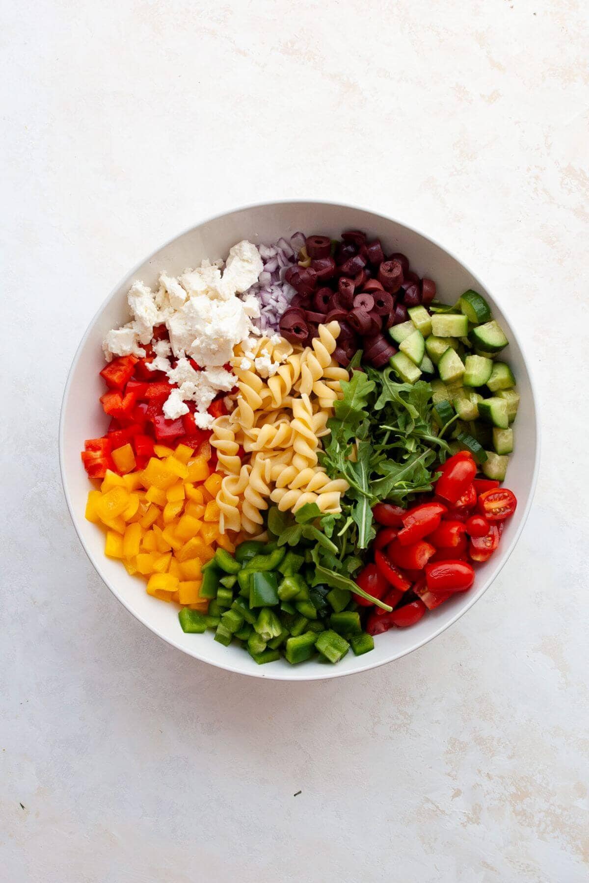 Chopped vegetables, arugula, olives, and feta arranged in a white bowl with cooked pasta before tossing.