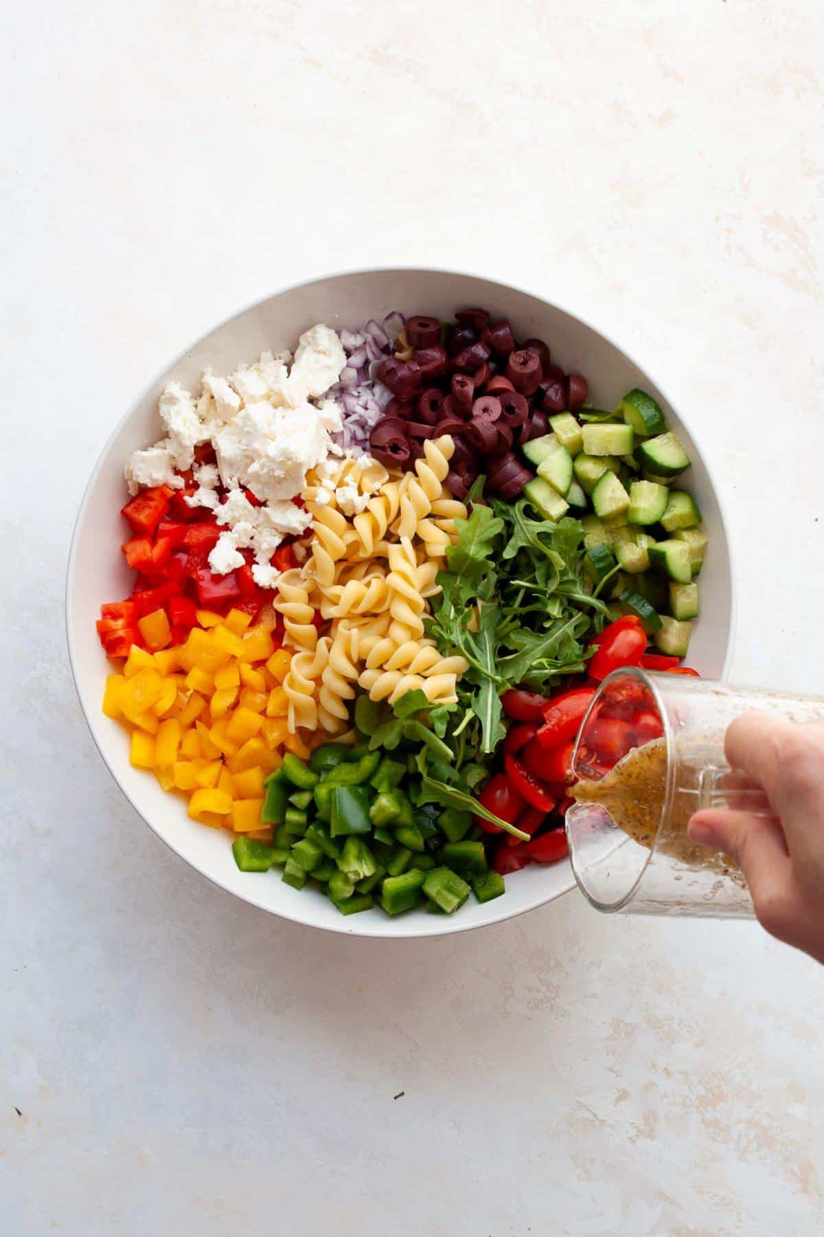 Homemade Greek dressing being poured over pasta salad ingredients in a white bowl.