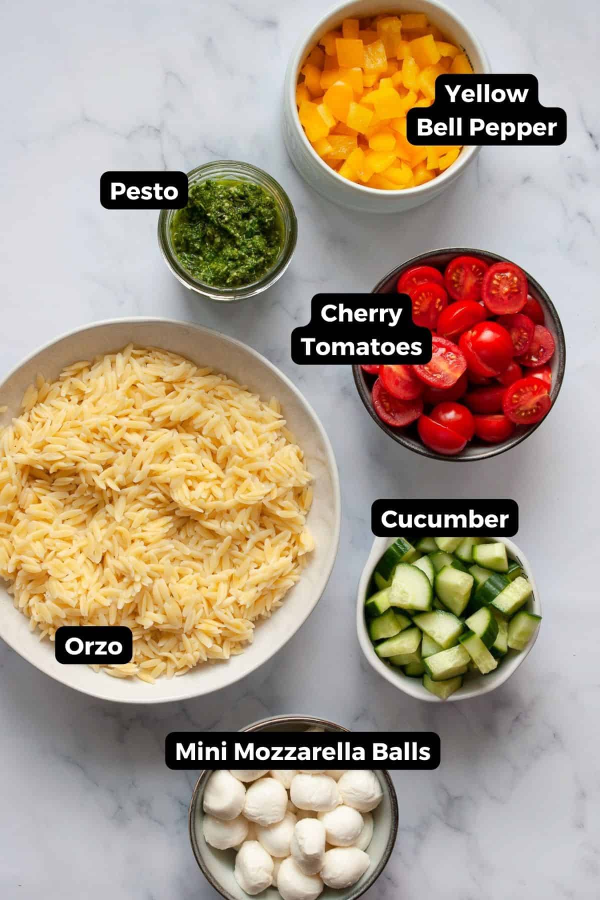 all ingredients in pesto orzo salad in bowls including cooked orzo, mini mozzarella balls, cherry tomatoes, pesto, yellow bell pepper, and cucumber