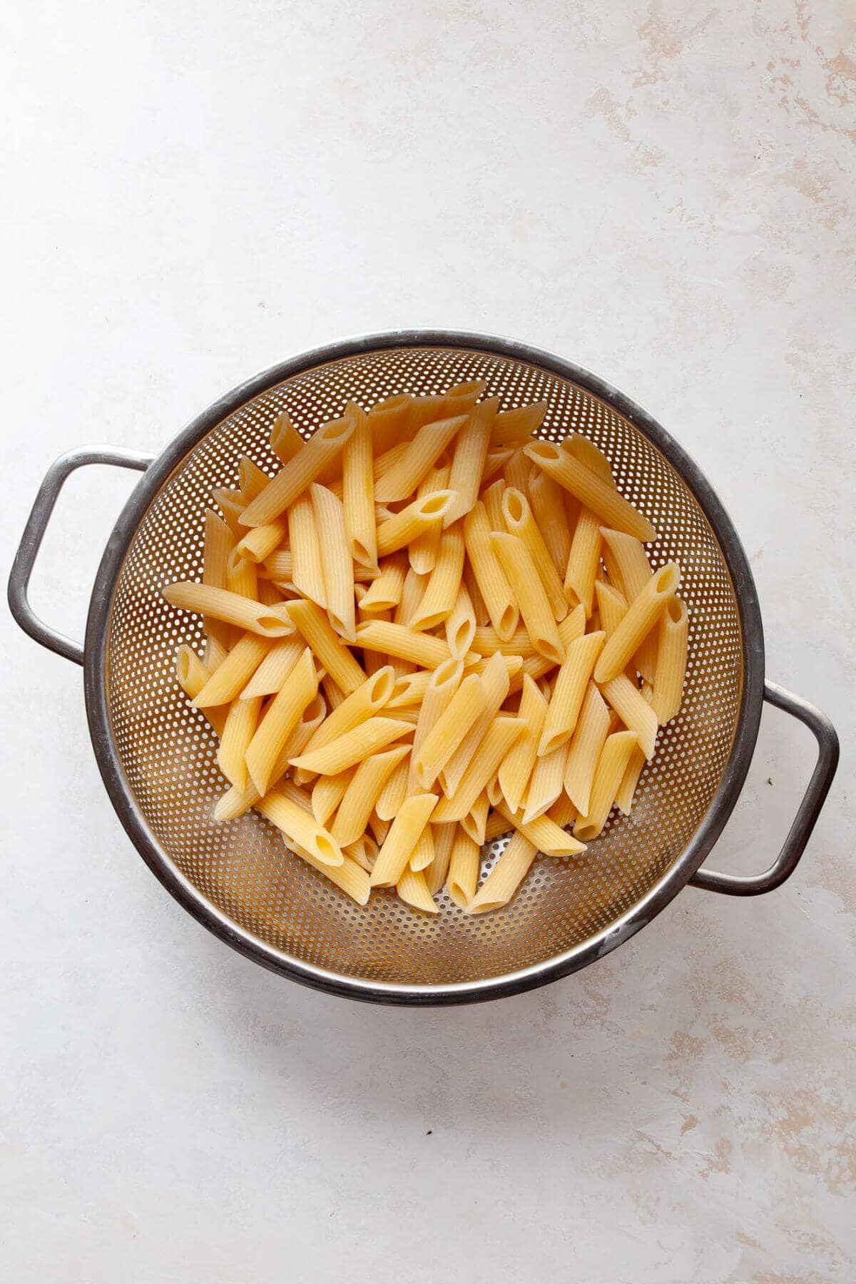 Cooked penne pasta being rinsed under cold water in a strainer to cool