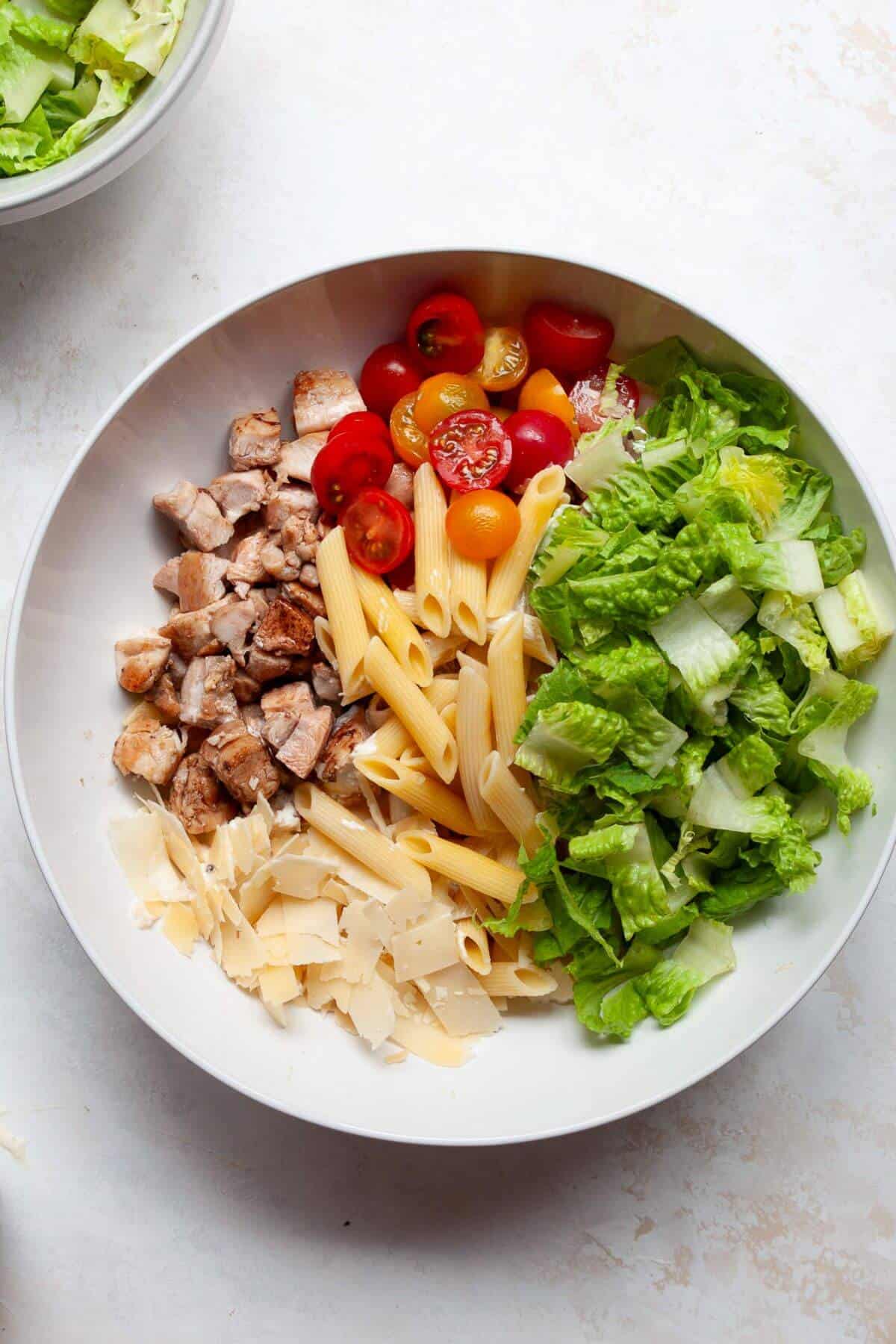 Large mixing bowl filled with chopped balsamic chicken, cherry tomatoes, romaine, parmesan, and pasta before tossing