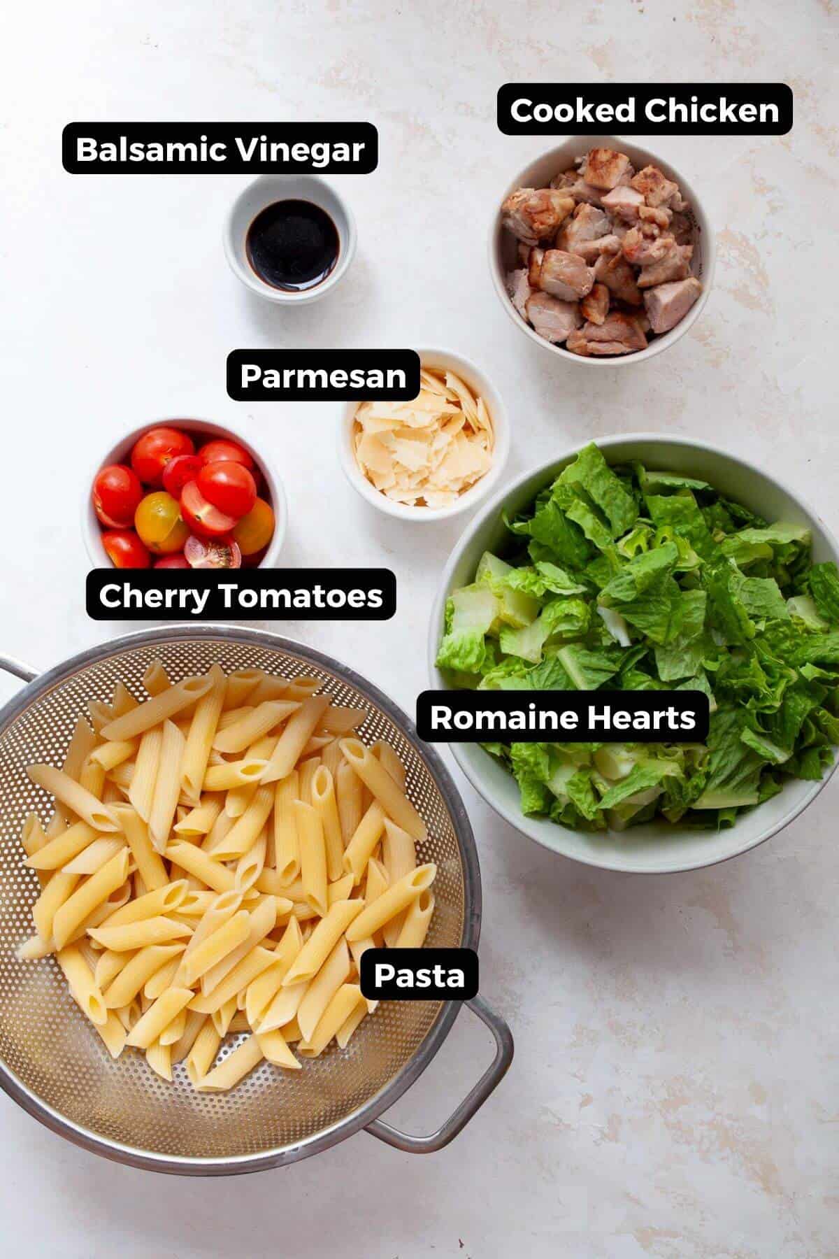 all ingredients in chicken Caesar pasta salad in bowls including pasta, lettuce, tomatoes, parmesan, chicken, and balsamic vinegar