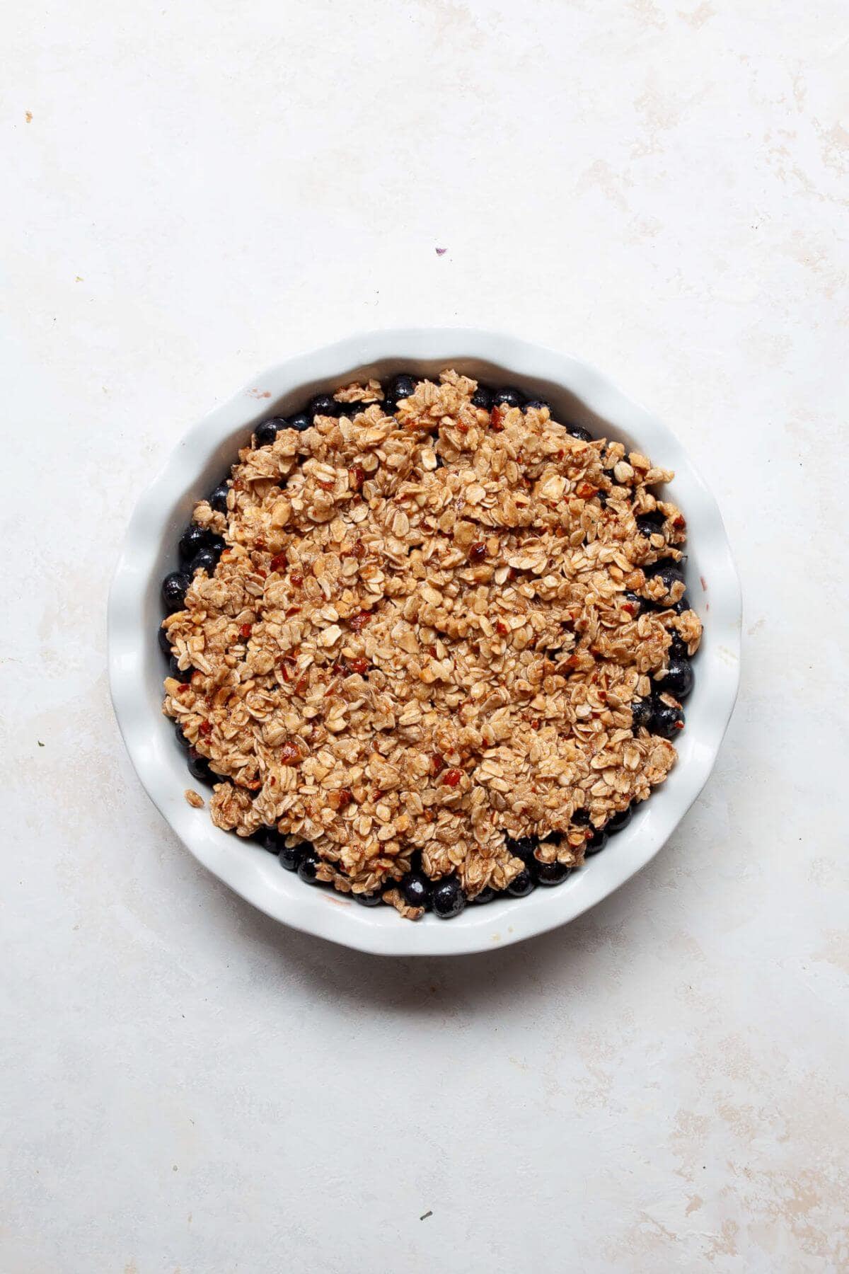 Almond-oat crumble topping sprinkled evenly over blueberry filling in a round baking dish before baking.