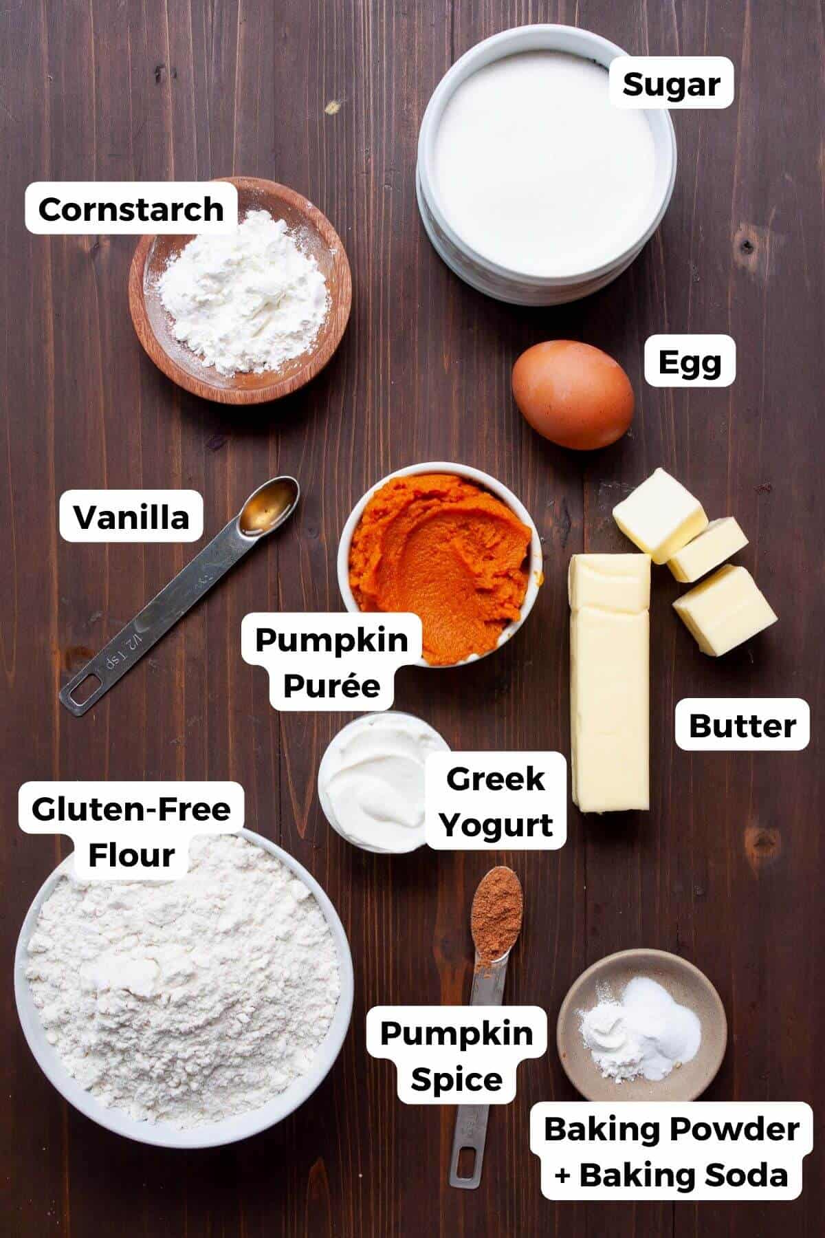Small bowls of ingredients for soft gluten-free pumpkin cookies with cream cheese frosting, including gluten-free flour blend, cornstarch, pumpkin purée, pumpkin spice, butter, egg, Greek yogurt, granulated sugar, brown sugar, baking powder, baking soda, cream cheese, maple syrup, vanilla extract, and salt, each labeled.