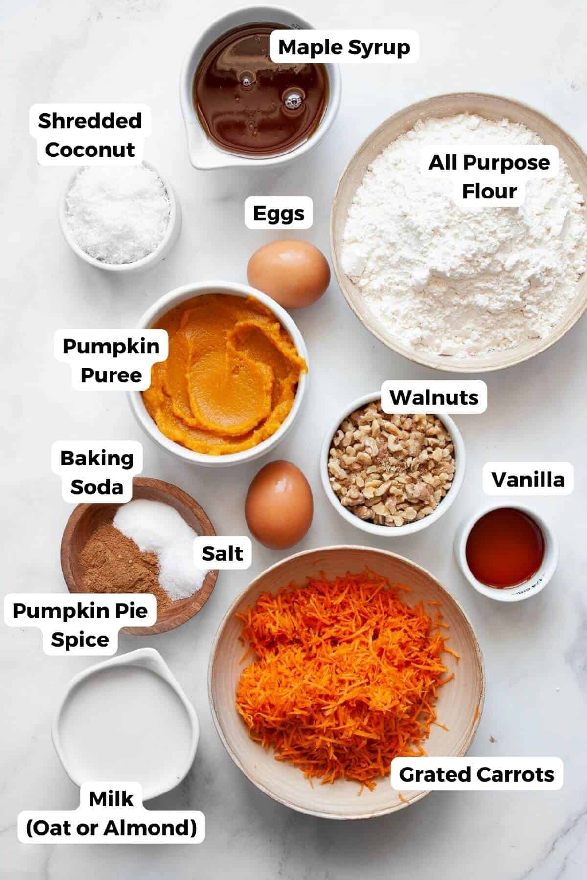 All ingredients for pumpkin carrot cake in small bowls with labels, including pumpkin purée, grated carrots, flour, maple syrup, coconut oil, eggs, walnuts, shredded coconut, and spices.