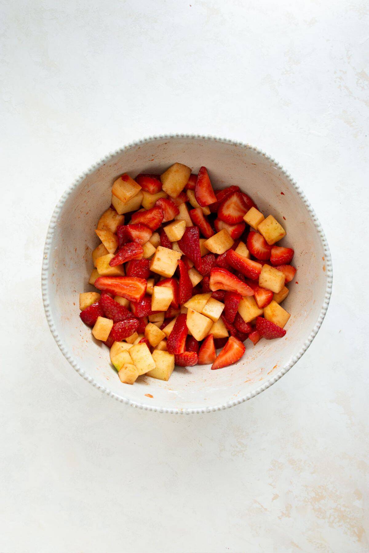 Chopped strawberries and apples tossed with starch and coconut sugar in a white mixing bowl.