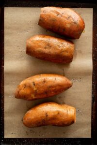 four raw sweet potatoes on a baking sweet with olive oil, salt, and pepper