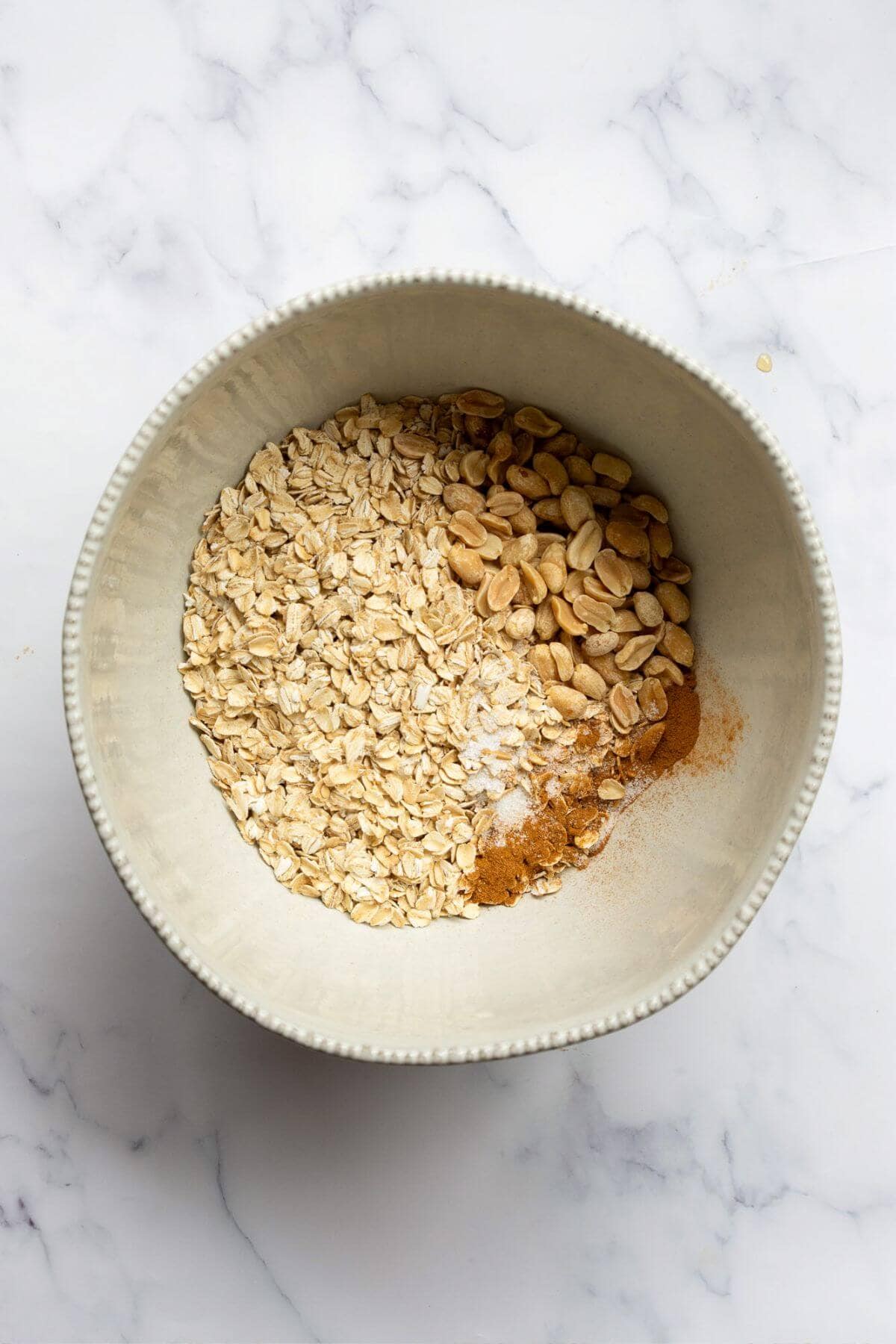 Rolled oats, roasted peanuts, cinnamon, and salt combined in a large mixing bowl on a marble counter.