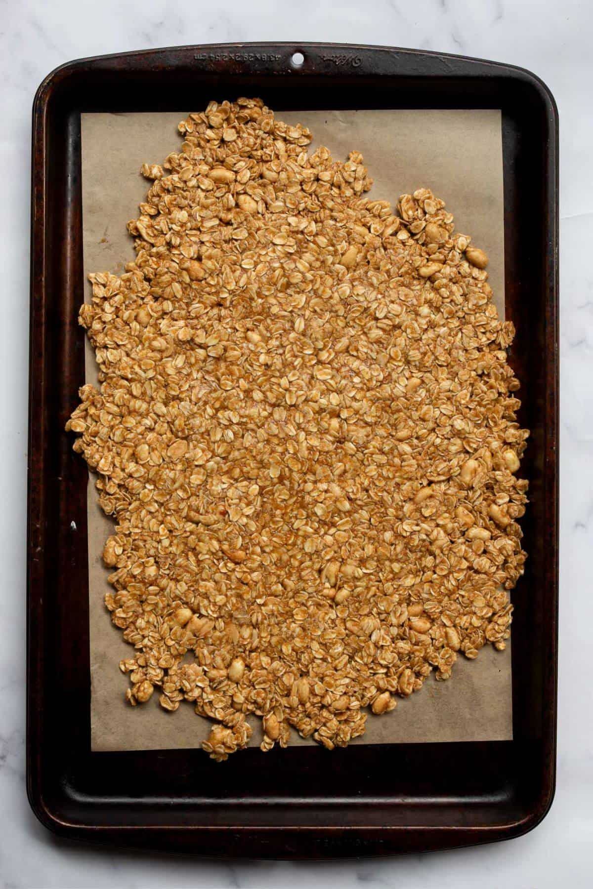 Unbaked peanut butter granola pressed into a flat, even layer on a parchment lined baking sheet, ready to bake at 325°F.