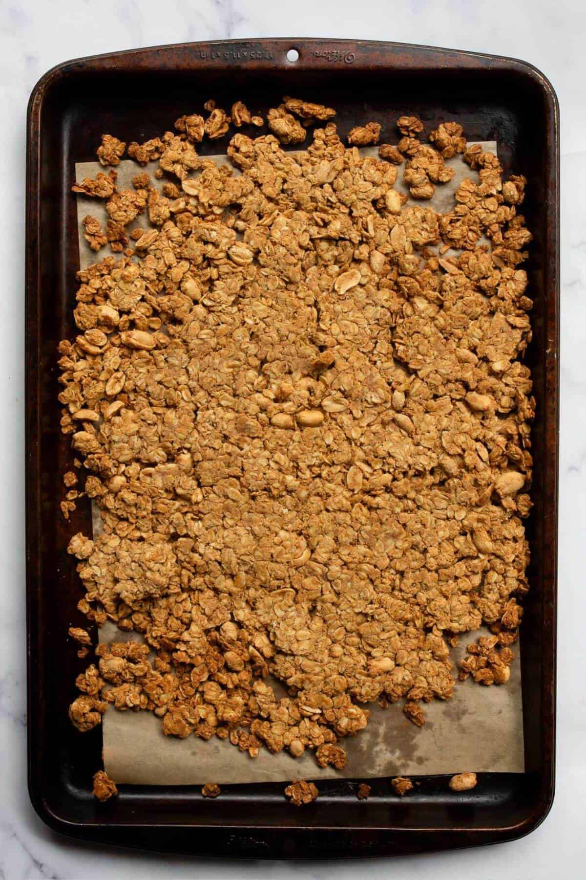 Golden brown peanut butter granola fresh out of the oven on a parchment lined baking sheet, ready to cool into chunky clusters.