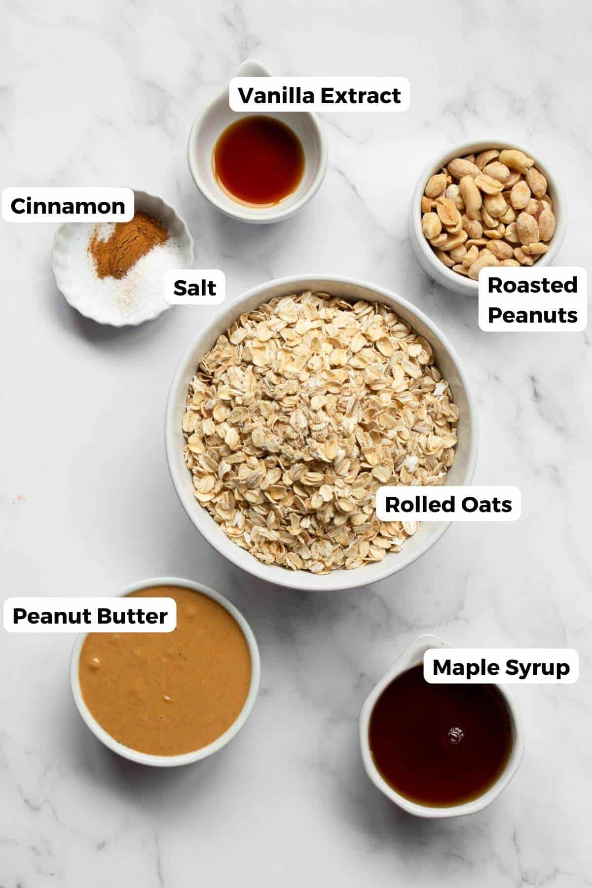 Ingredients for healthy peanut butter granola laid out on a marble countertop, including rolled oats, natural peanut butter, maple syrup, vanilla extract, cinnamon, salt, and roasted peanuts.