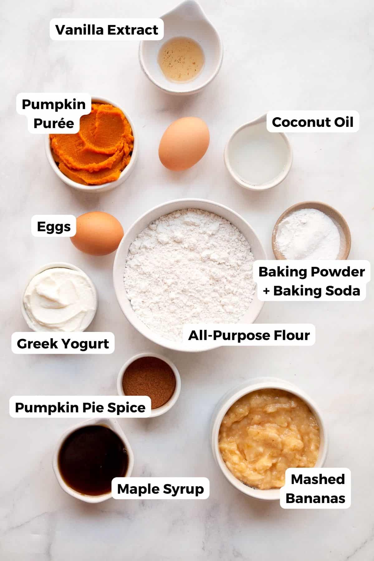 All the ingredients for healthy banana pumpkin muffins in small bowls with labels - ripe bananas, pumpkin purée, maple syrup, Greek yogurt, eggs, flour, baking powder, baking soda, pumpkin pie spice, cinnamon, and salt.