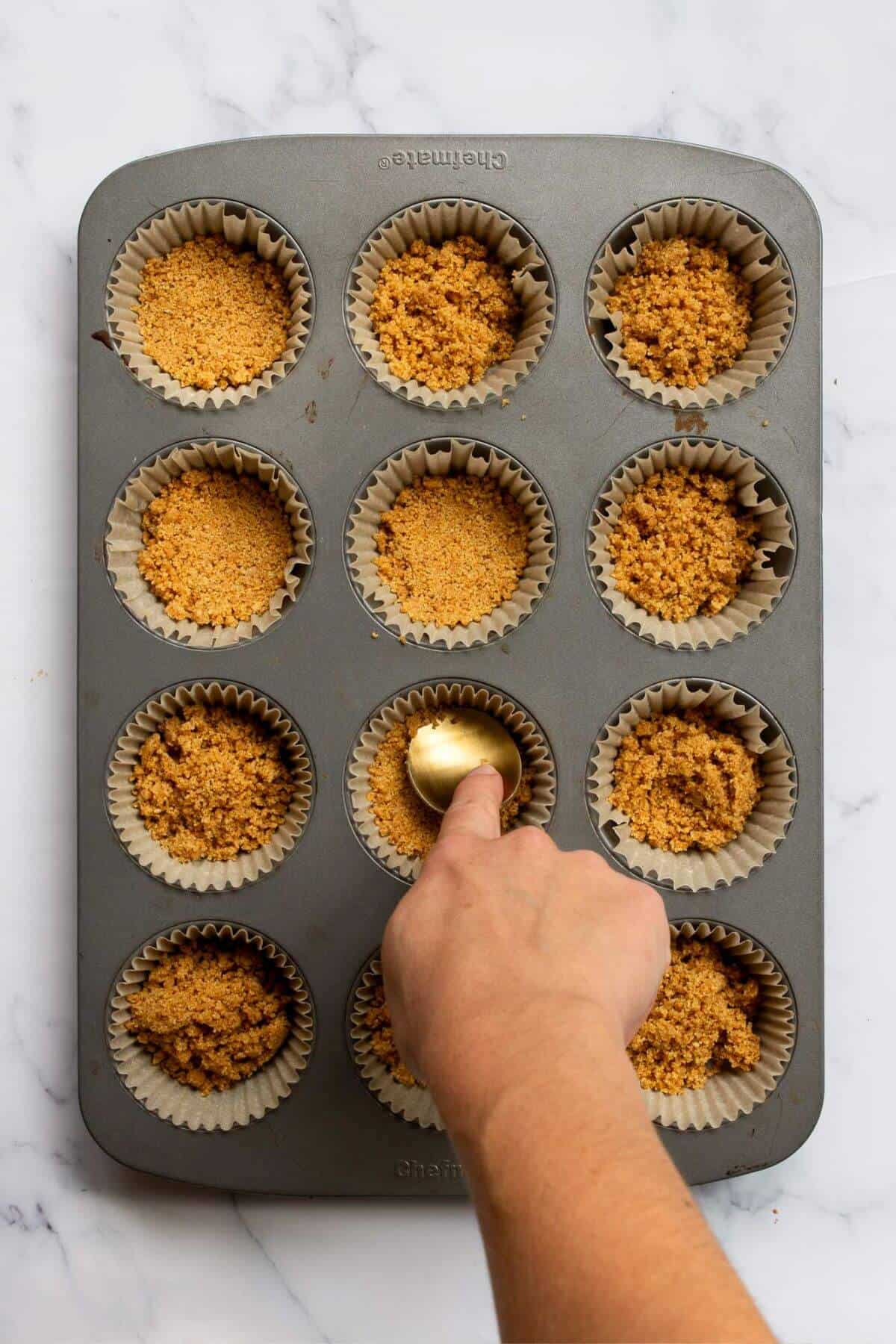 Using a spoon to press graham cracker crust into muffin cup liners for mini pumpkin cheesecakes.